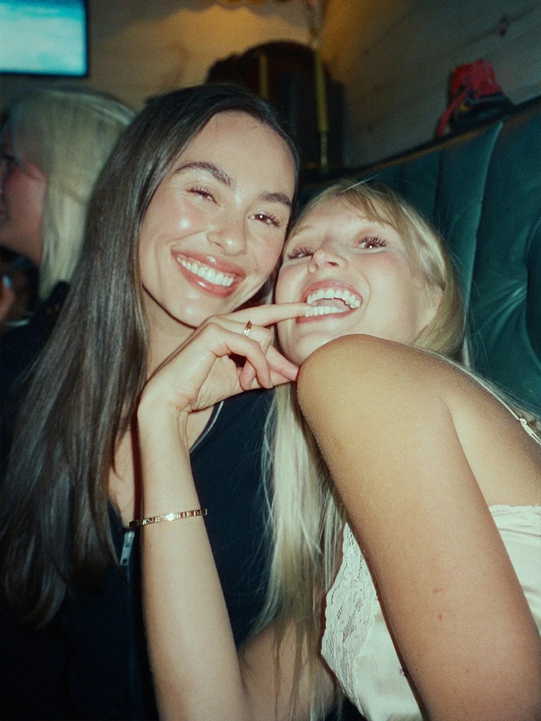 Two young women smiling and laughing together in a social setting, with one touching the other's face.