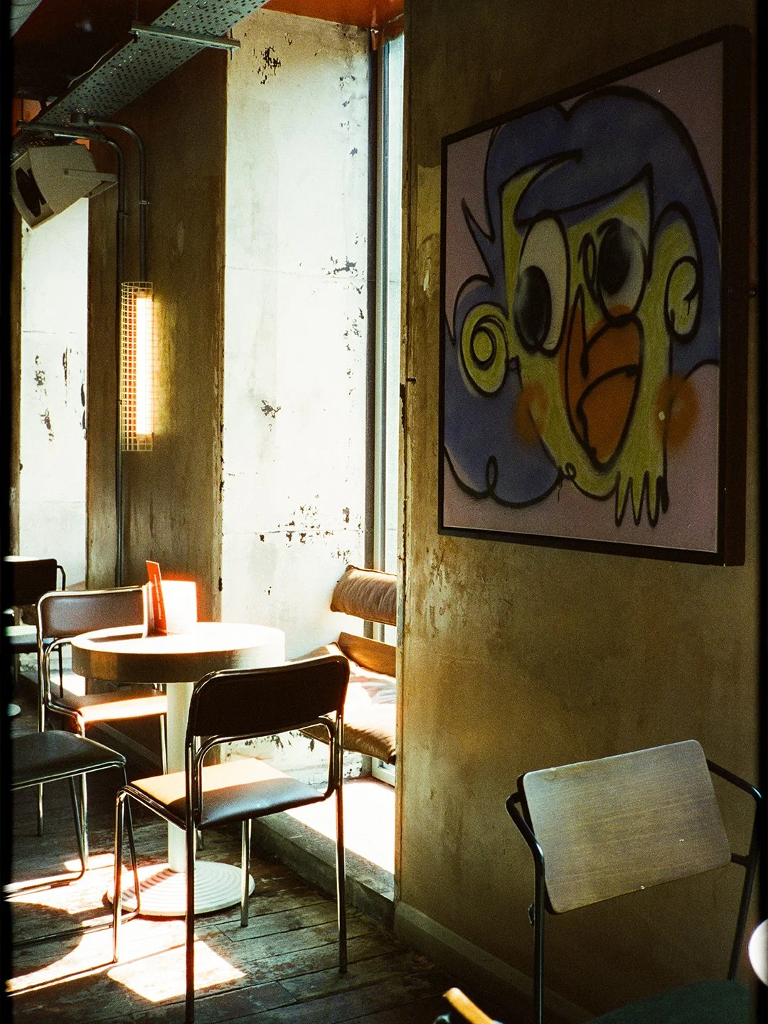 Interior of a cafe with a round table, black chairs, a bench, a colorful cartoon-style bird painting on the wall, and sunlight streaming through a window.