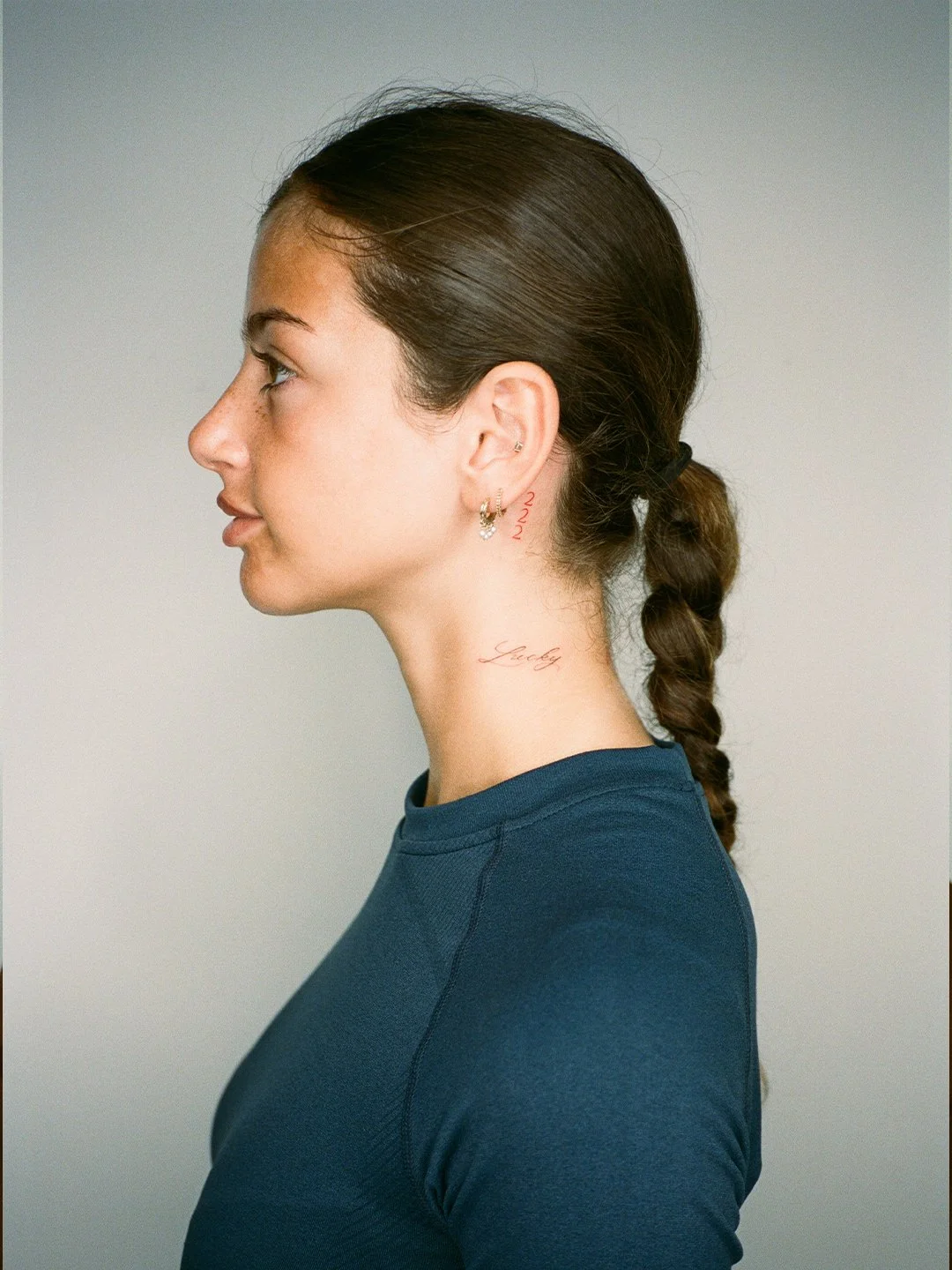 Profile view of a woman with brown hair tied in a braid, wearing a blue shirt, with small earrings and tattoos on her neck and behind her ear.
