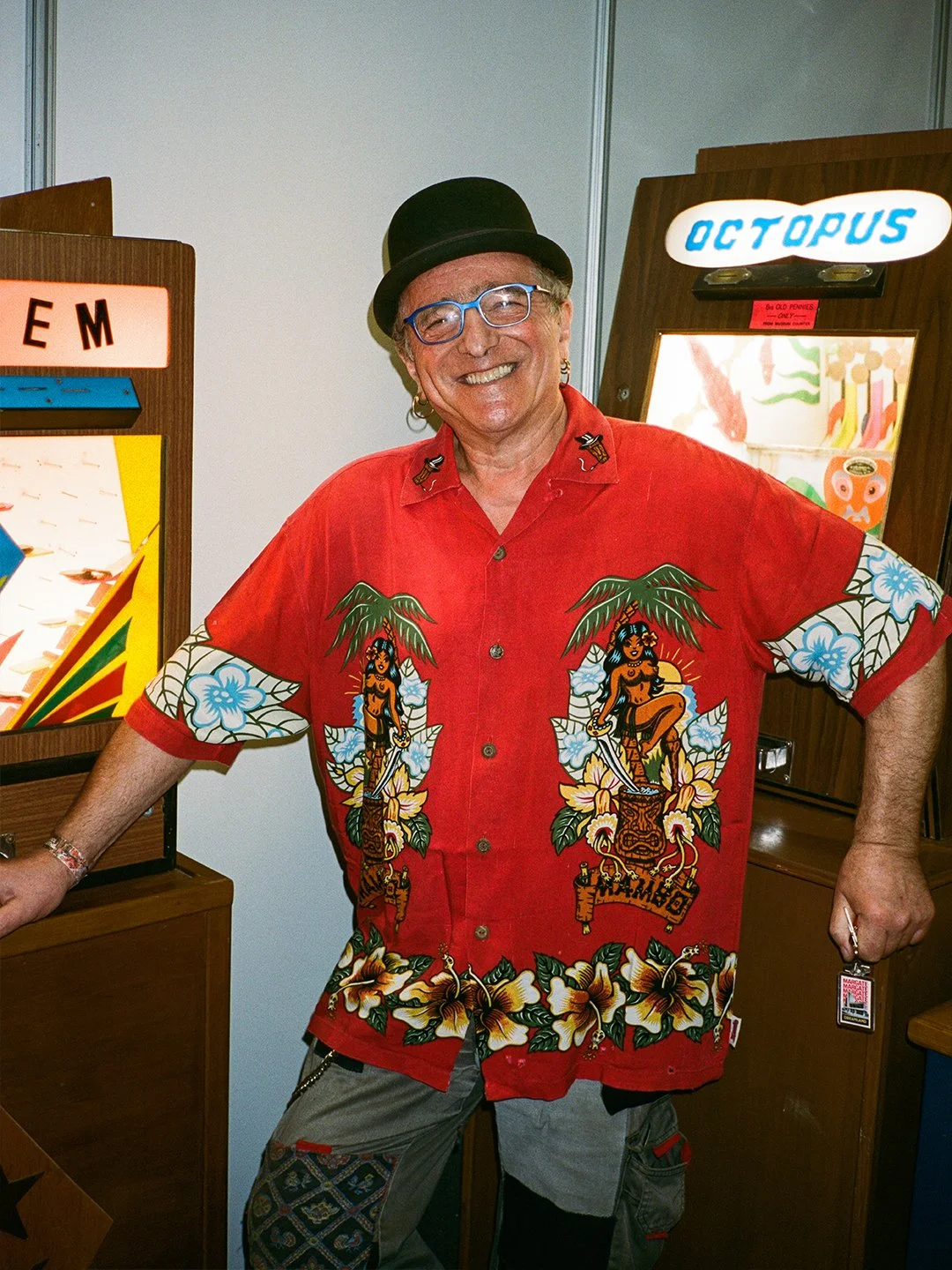 A man wearing a black hat, blue glasses, and a red Hawaiian shirt with tropical and tribal designs, posing in front of pinball machines at an arcade.