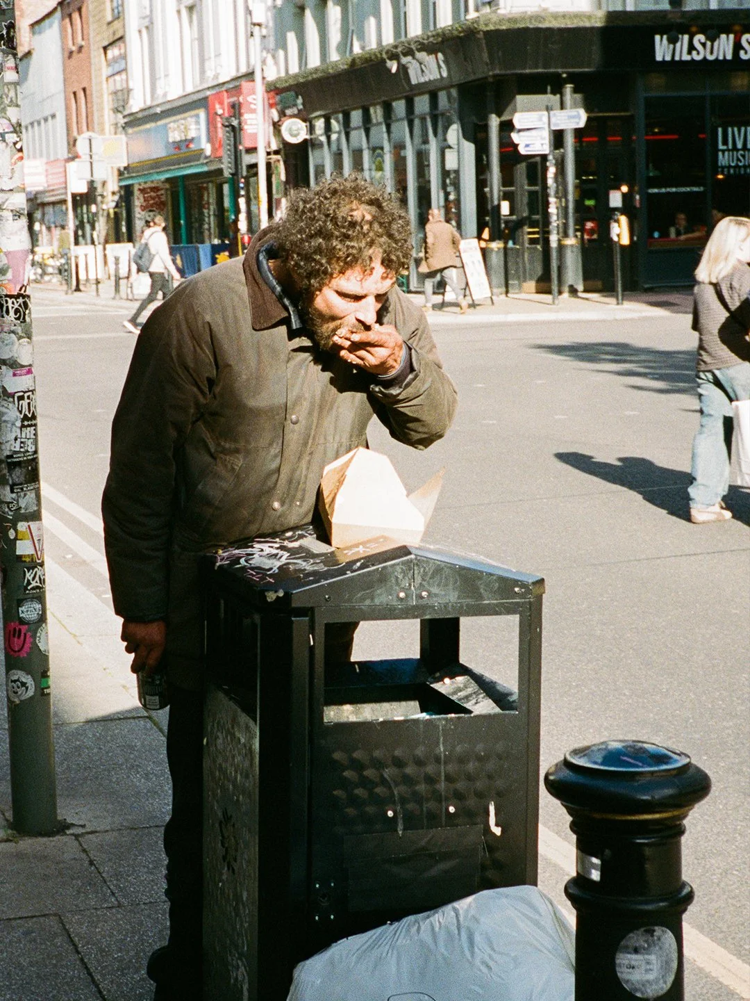 A man with curly hair in a brown jacket is eating something from his hand on a city sidewalk. There is a black trash can in front of him with some paper or food container on top, and a white bag or blanket on the ground at its base. People are walkin