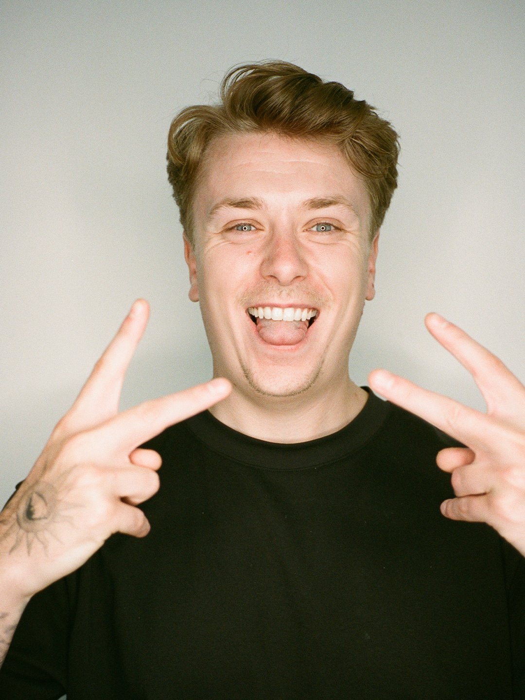 A young man with light brown hair smiling widely, showing his teeth, and making peace signs with both hands. He is wearing a black shirt, and the background is plain. His expression is joyful and energetic.
