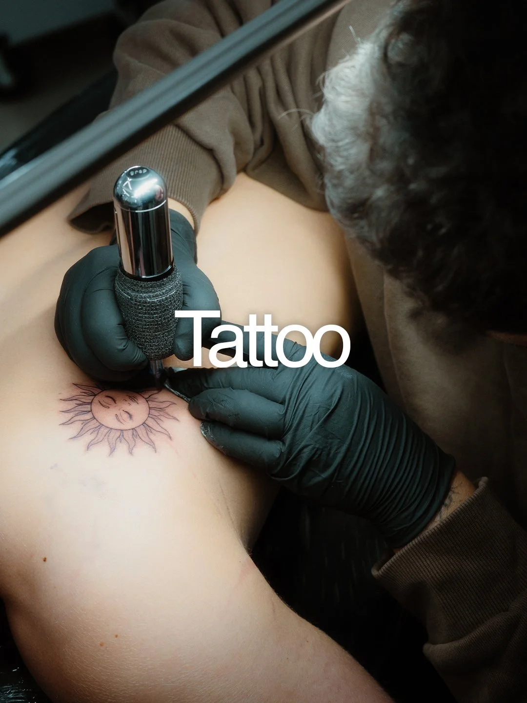 A person with black gloves tattoos a sun with a face on someone's shoulder using a tattoo machine.