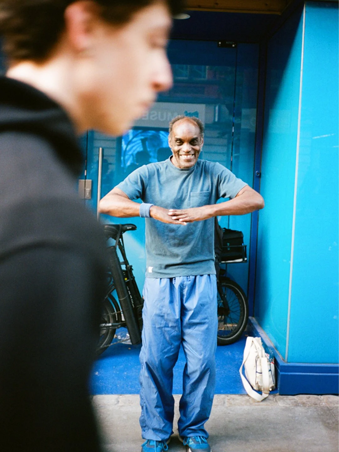 A smiling man in a gray T-shirt and blue athletic pants stands with arms crossed in front of a blue wall, with a bicycle and a white bag on the ground beside him. In the foreground, a blurred profile of a younger person is visible.