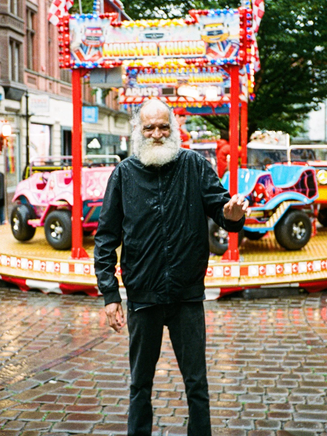 An elderly man with a white beard and wet black raincoat standing in front of a colorful outdoor amusement ride with toy trucks, on a rainy day