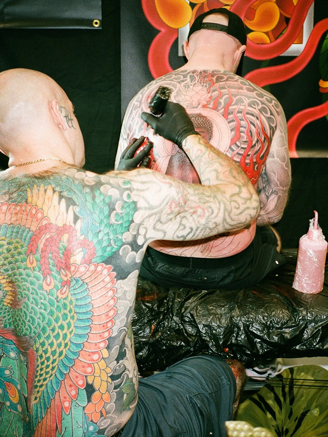 A tattoo artist is tattooing a large, colorful design on a man's upper back. The artist has numerous tattoos on his arms and is wearing black gloves. The man receiving the tattoo is sitting and wearing a black cap backward, and their head is bowed fo