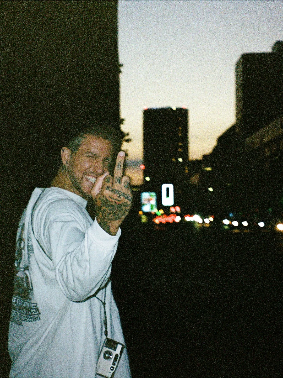 A young man with tattoos on his hand and face, smiling, making a peace sign, standing in front of a city street at dusk, with high-rise buildings and blurred car lights in the background.