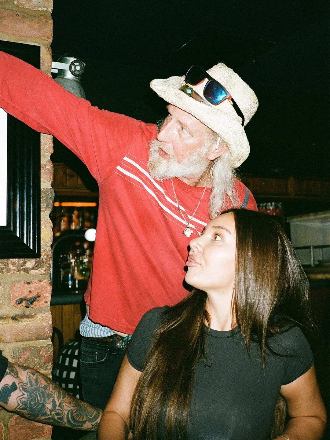A man with long gray hair and a beard, wearing a straw hat with sunglasses on top, pointing at a picture on the wall, while a young woman with long brown hair sitting beside him looks up at him in interest.