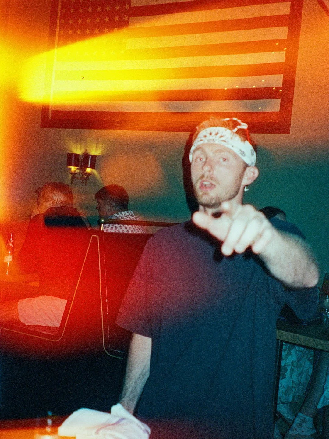 A man wearing a white bandana pointing towards the camera in a room with an American flag on the wall.