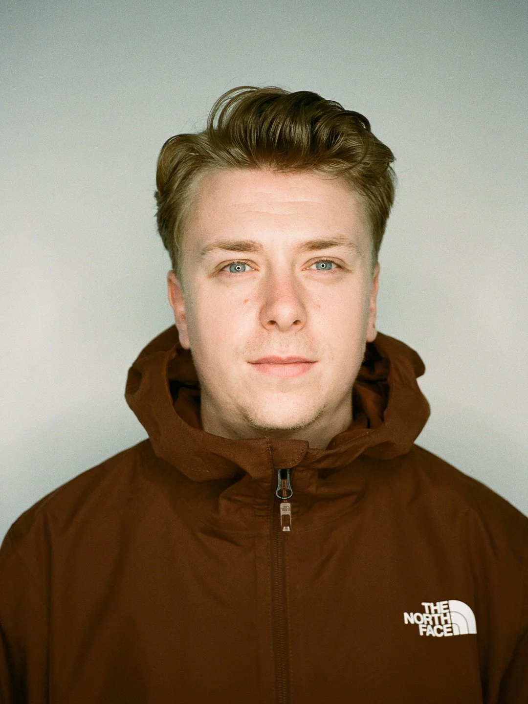 Headshot of a young man with light skin, blue eyes, and short brown hair, wearing a brown The North Face jacket against a plain light background.