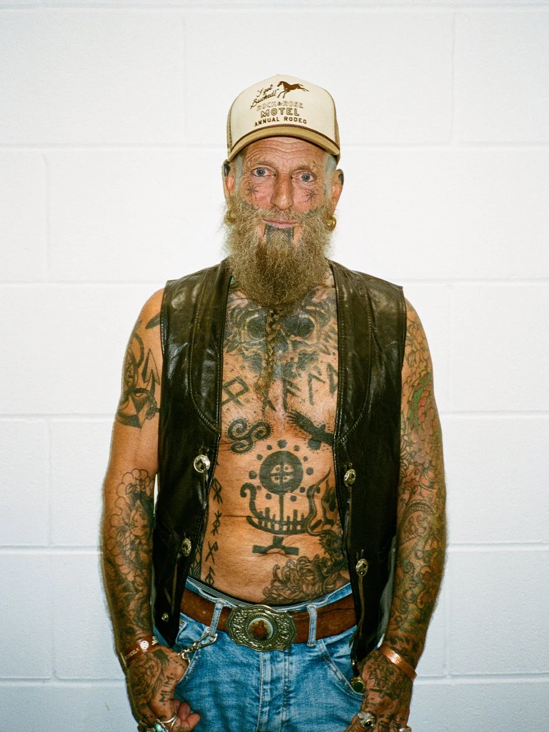 Man with tattoos on arms and chest, beard, wearing a baseball cap, black leather vest, jeans, and a large belt buckle.