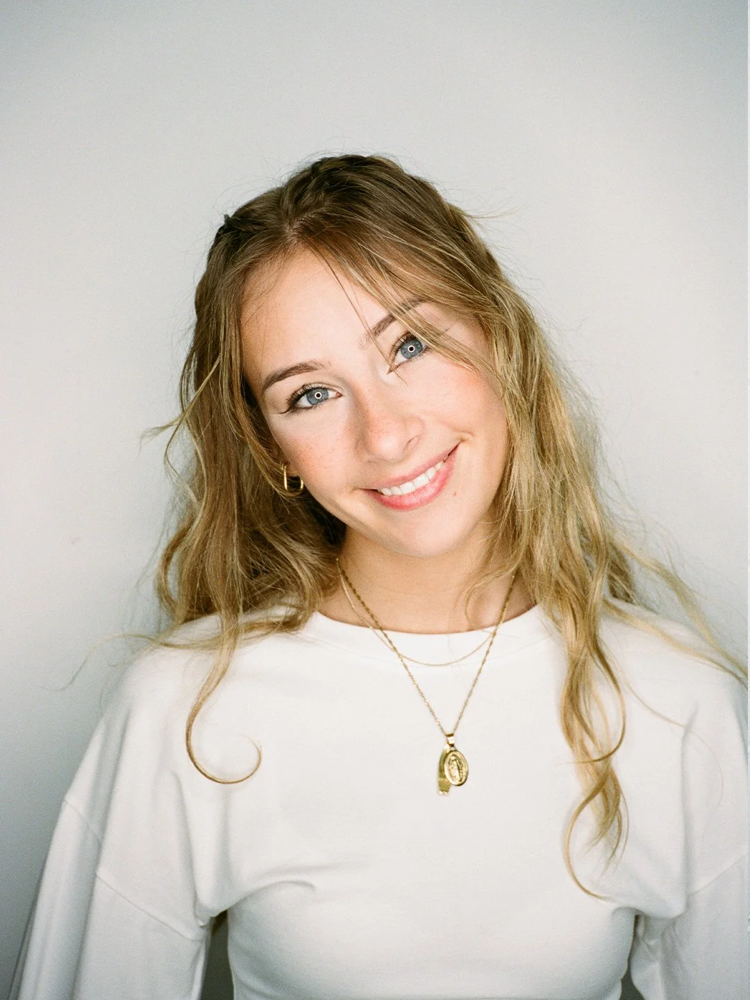 A woman with wavy blonde hair and blue eyes smiling, wearing a white top and gold jewelry, in front of a plain white background.