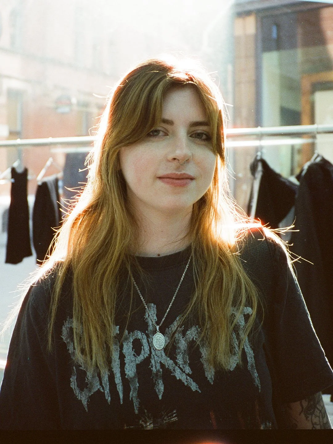 A woman with long wavy hair wearing a black graphic T-shirt and a silver necklace, standing outdoors near a clothing rack with black garments, with sunlight illuminating her hair.