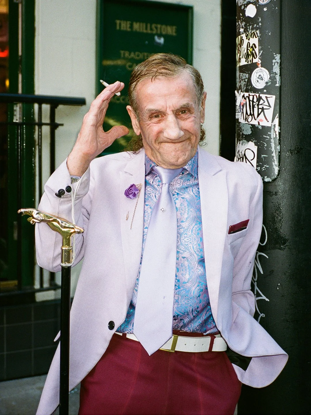 Older man with slicked back hair wearing a white blazer, patterned blue shirt, lavender tie, burgundy pants, and white belt, with a purple flower lapel pin, standing outside a bar or club, smiling and raising his right hand to his head, beside a blac
