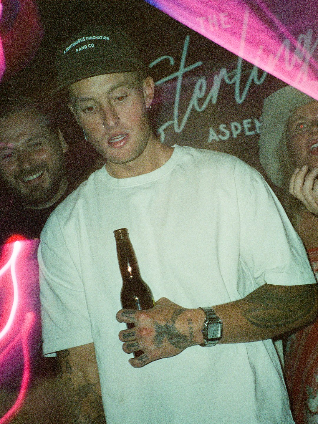 Person holding a beer bottle, with tattoos on hand, wearing a white t-shirt, black cap, and watch, in a dimly lit party setting with signs that read 'The Vintage Aspen'.