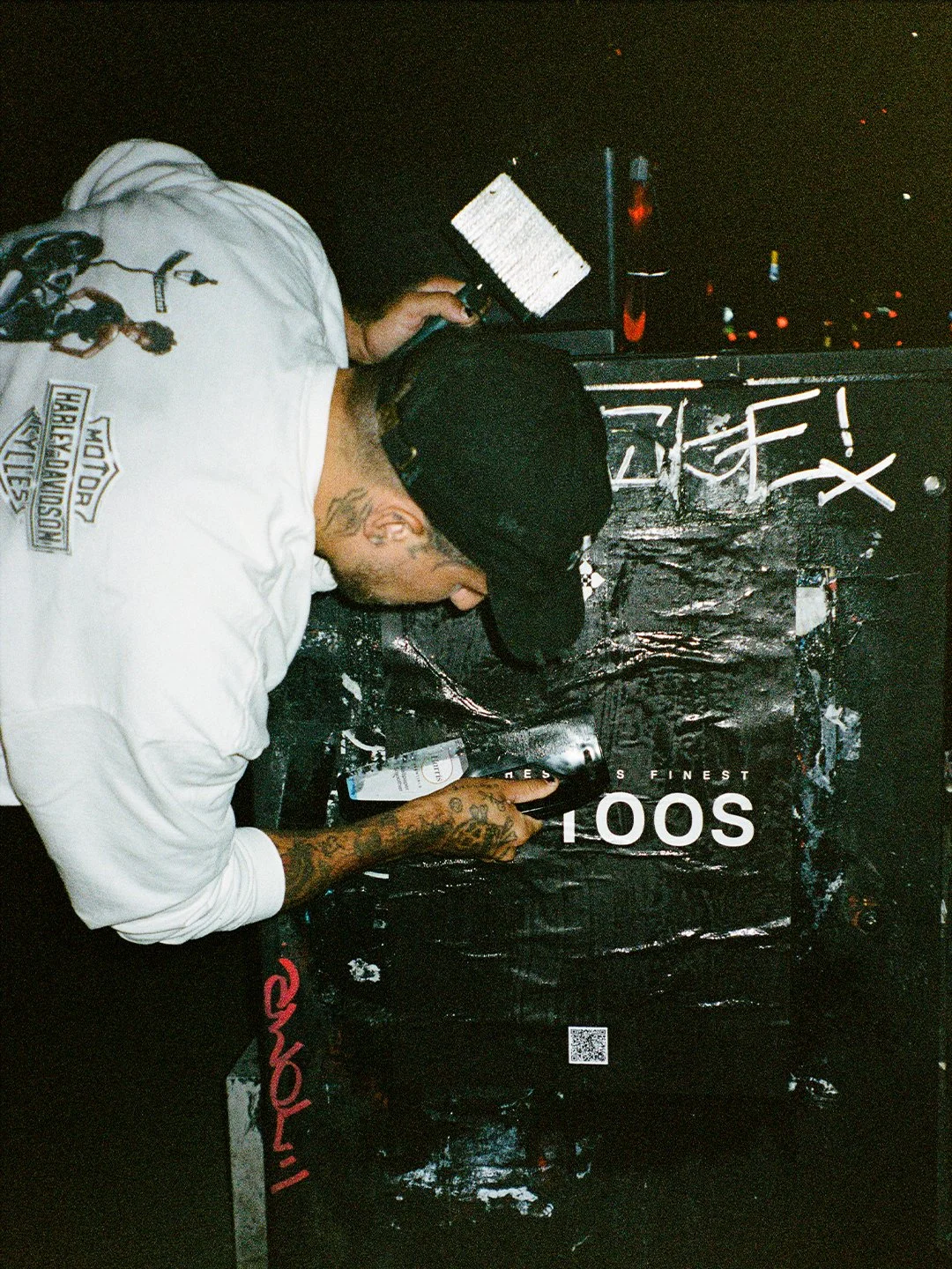 A man with tattoos wearing a white Harley-Davidson hoodie and a black cap is in a dark room, leaning over a black surface, writing or drawing with a marker, next to a partially visible laptop and graffiti on the wall.