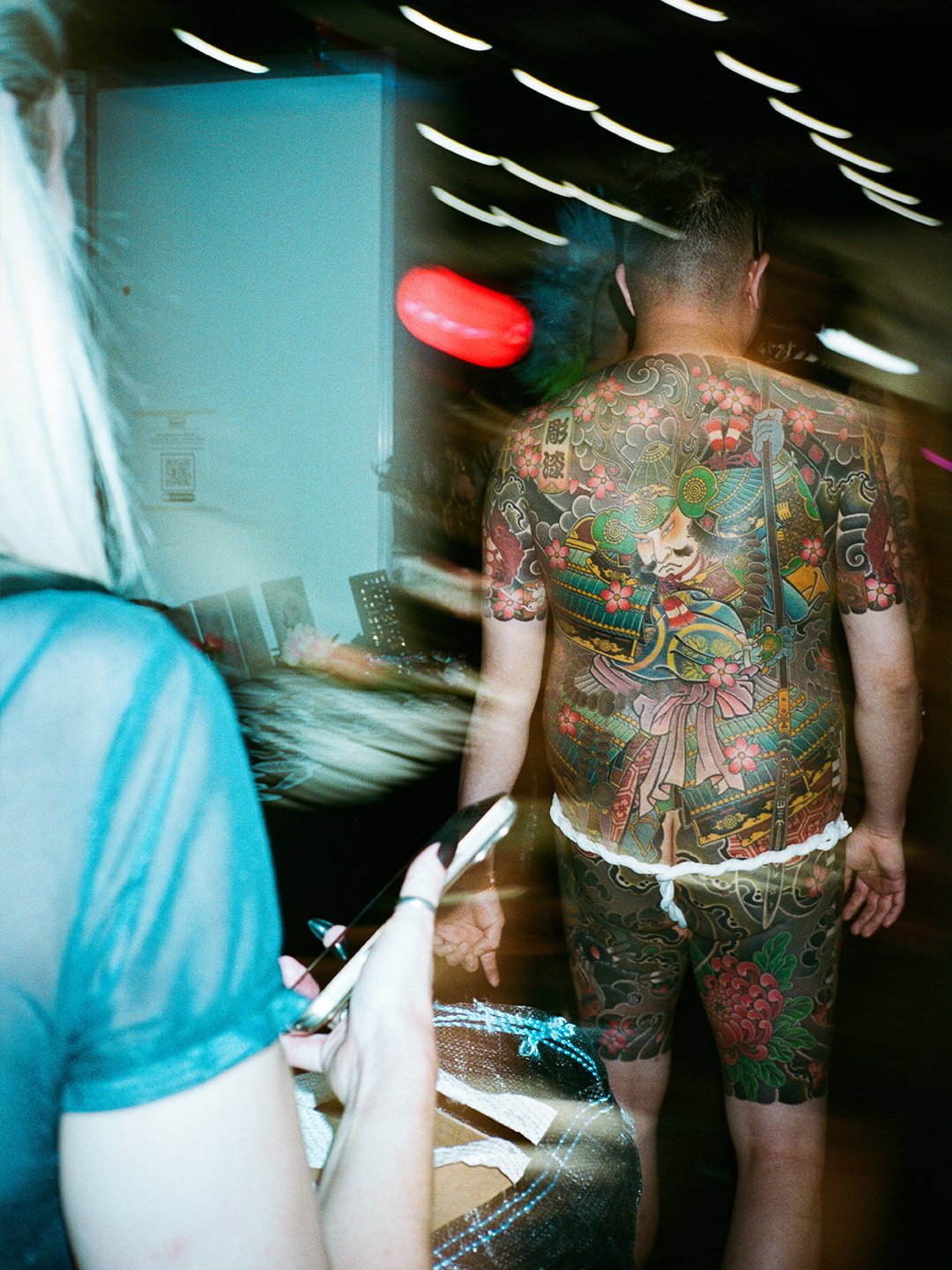 Back view of a person with a large colorful Japanese-style tattoo on their back, wearing shorts, in a dimly lit indoor setting with motion blur and streaks of light.