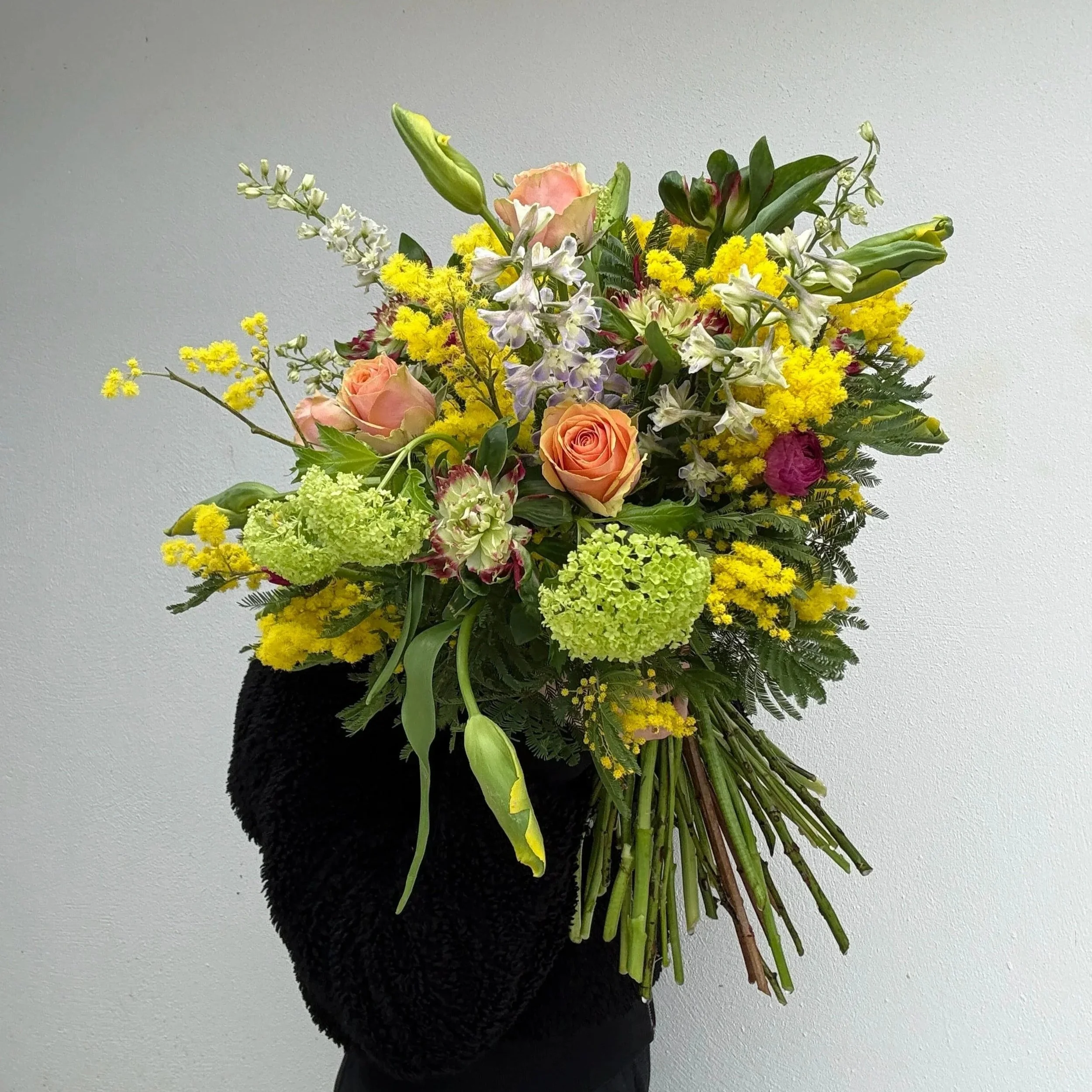 Person wearing black gloves holding a colorful bouquet of flowers against a plain white wall.