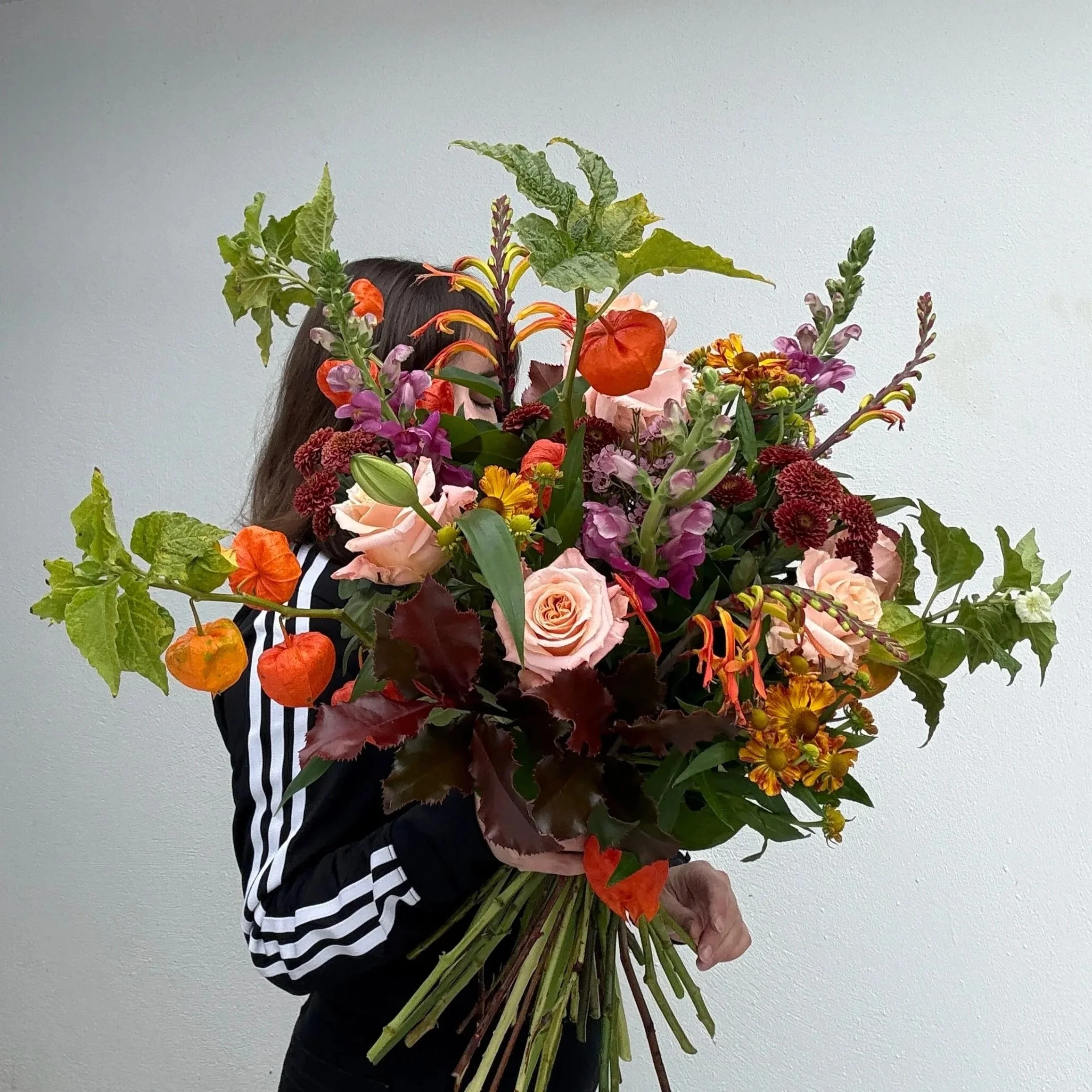 Person holding a large, colorful bouquet of various flowers including roses, lilies, and other blooms, partially obscuring their face.