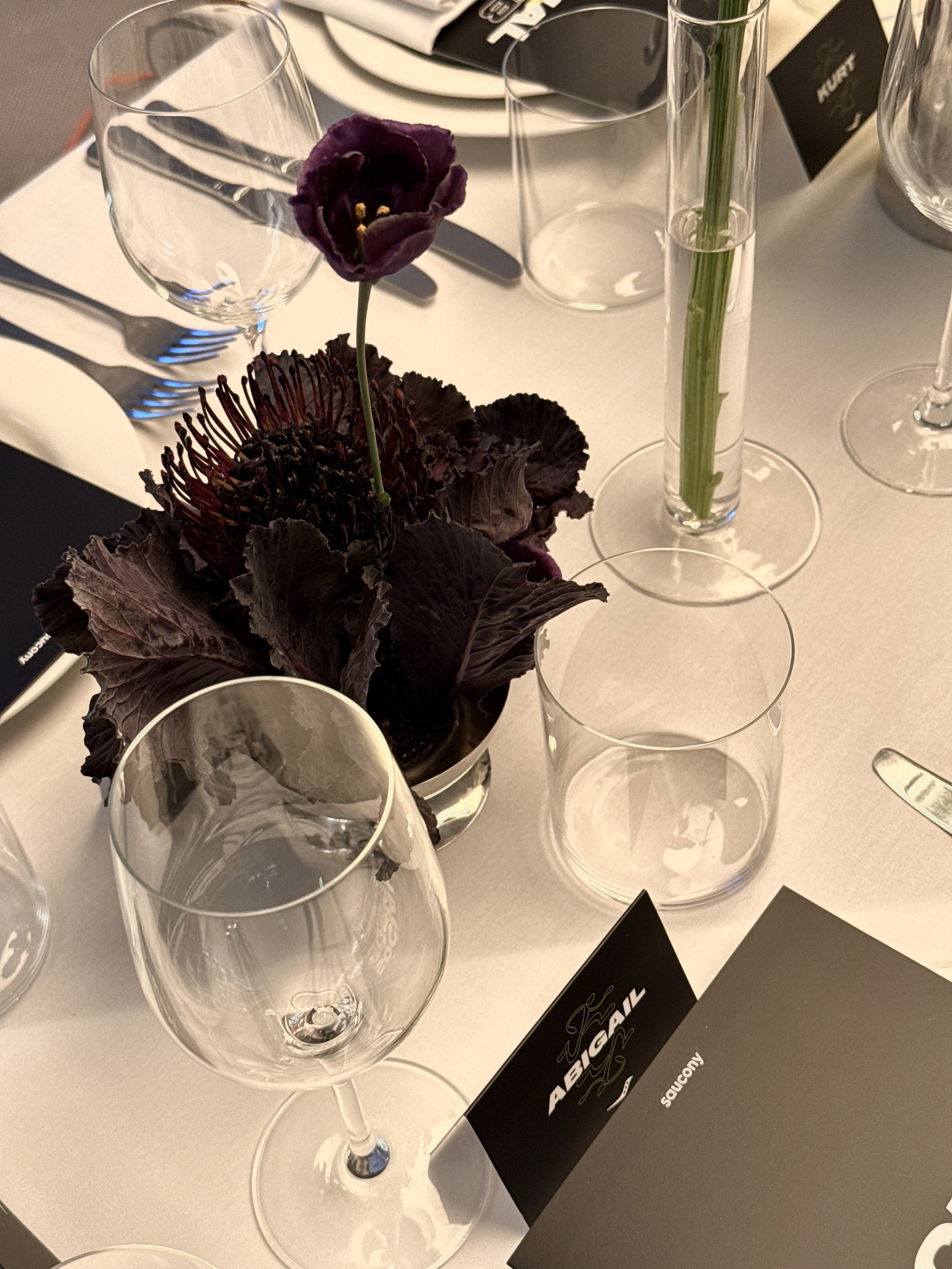 A table setting with empty wine glasses, a tall vase with a single purple flower and dark purple foliage, a black and white name card, and a partially visible menu or program, at a formal or semi-formal event.