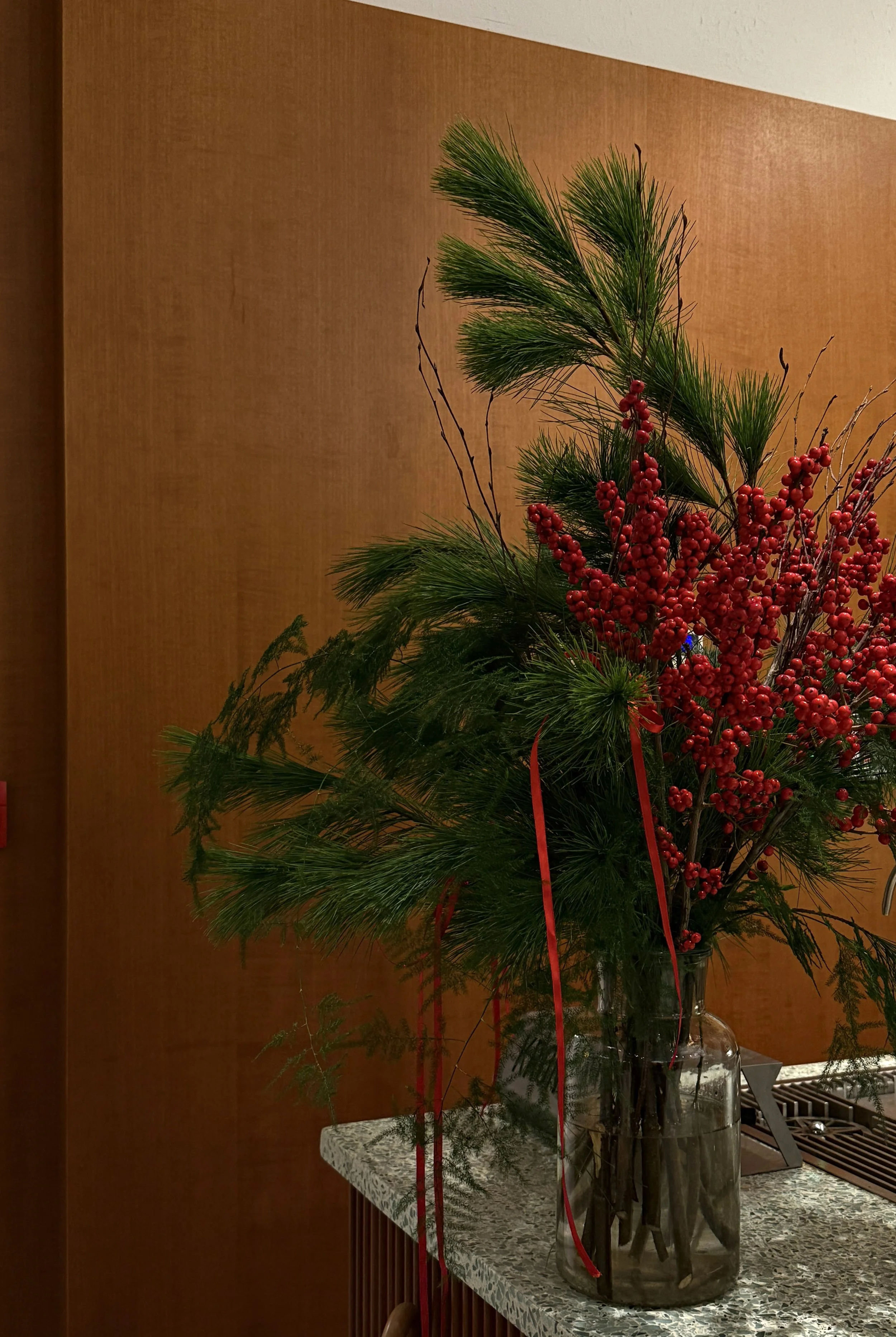 A flower arrangement with pine branches and red berries in a clear glass vase on a marble table against a wooden wall.