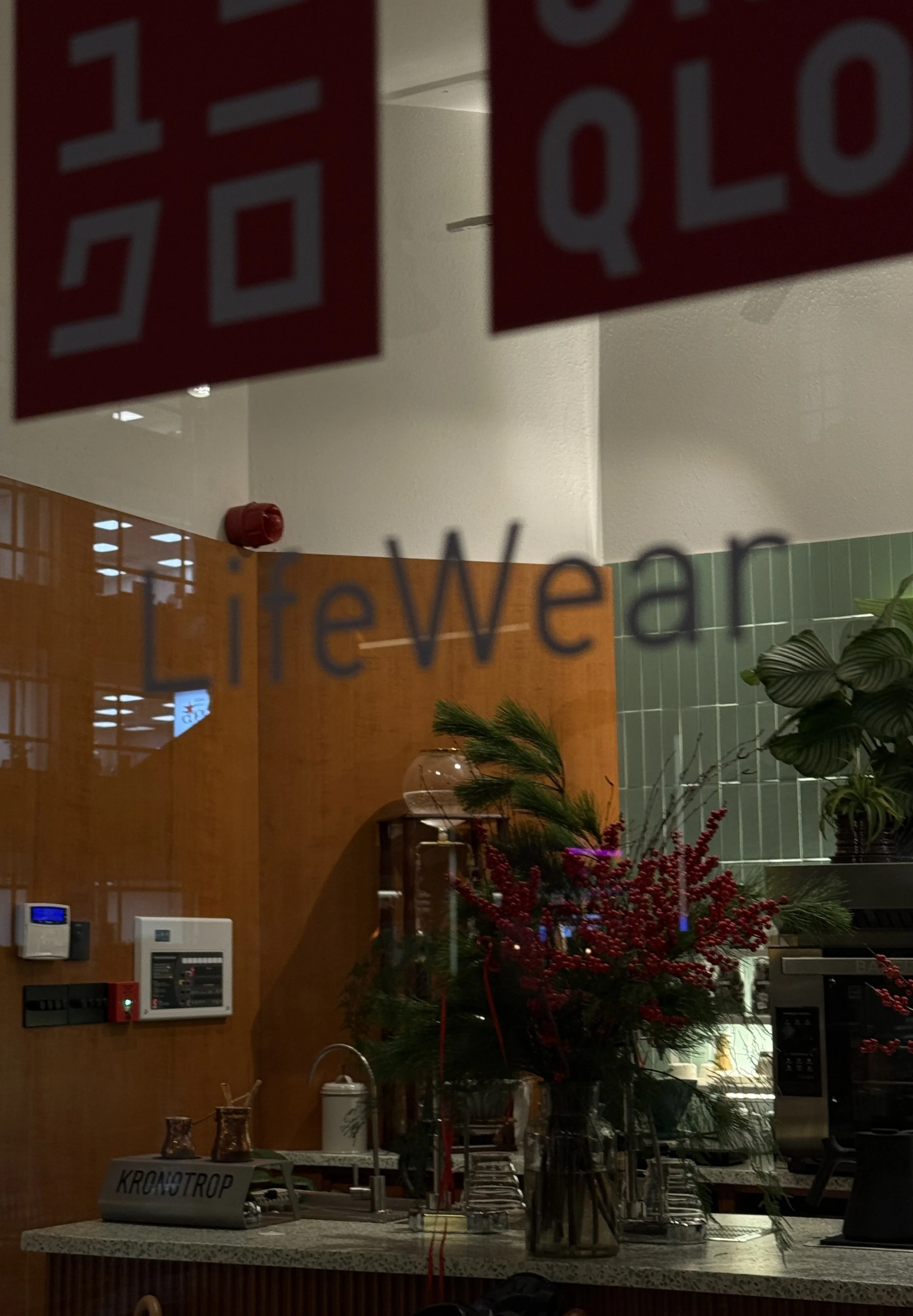 View of an indoor kitchen area through a glass door with the word 'LifeWear' written on it. Inside, there is a floral arrangement with red berries and green leaves on a counter, along with various kitchen appliances and a tiled wall.