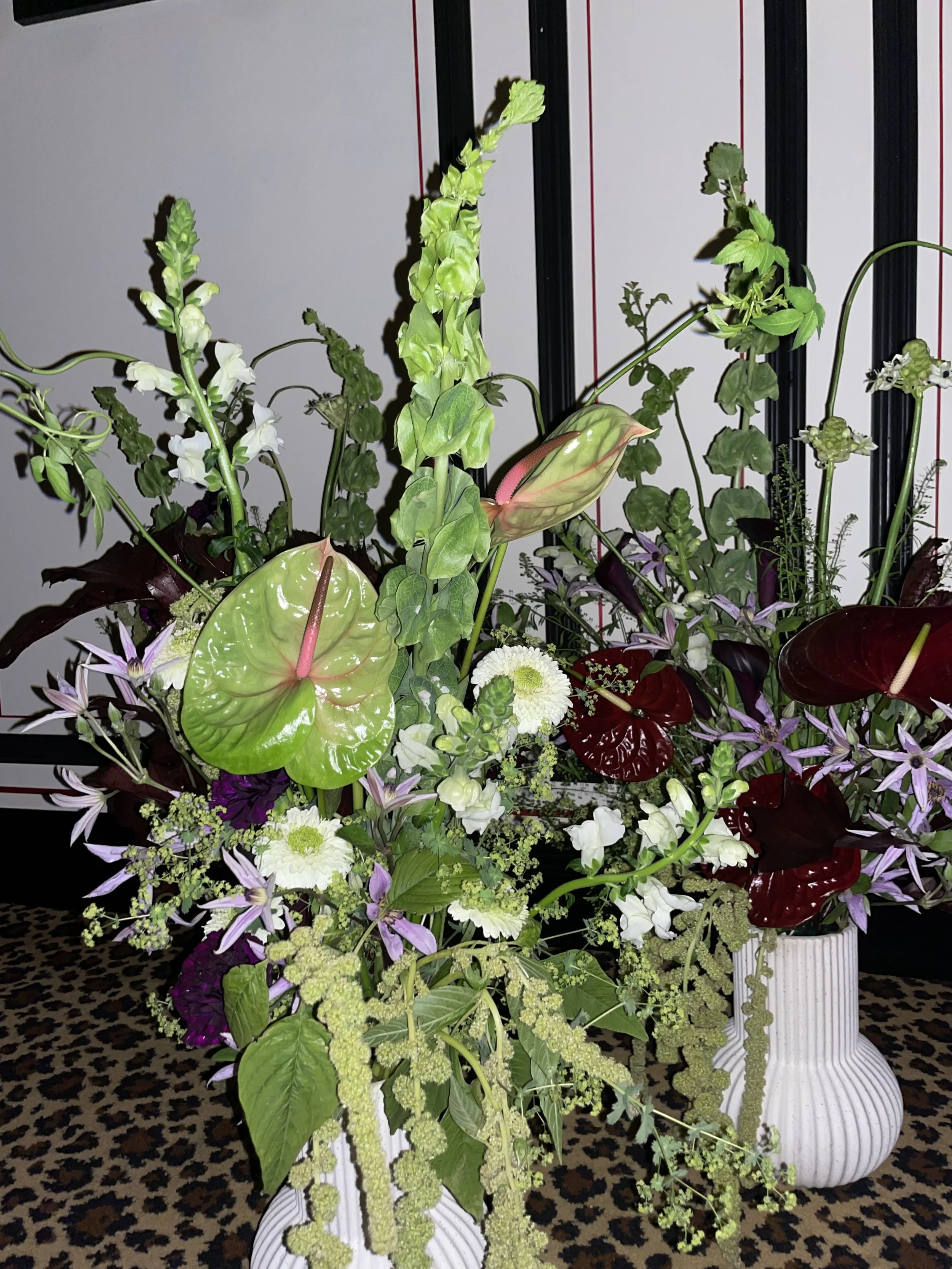 Two large white vases with colorful floral arrangements of a variety of green, purple, white, and red flowers and plants on a leopard print surface.
