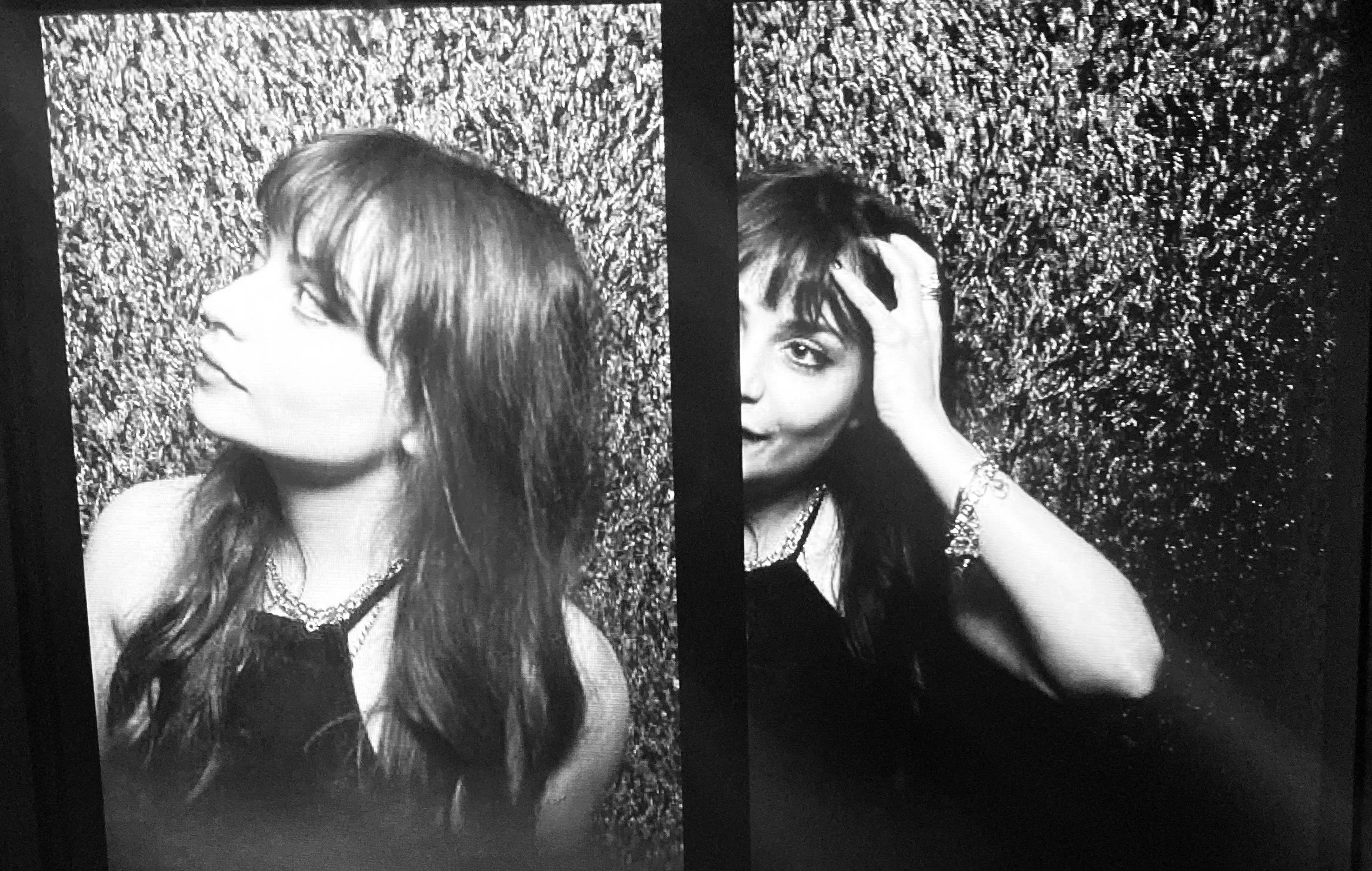 Black and white photo of two women with dark hair posing against a textured wall. The woman on the left is looking to the side, while the woman on the right is looking at the camera with her hand on her forehead, smiling.