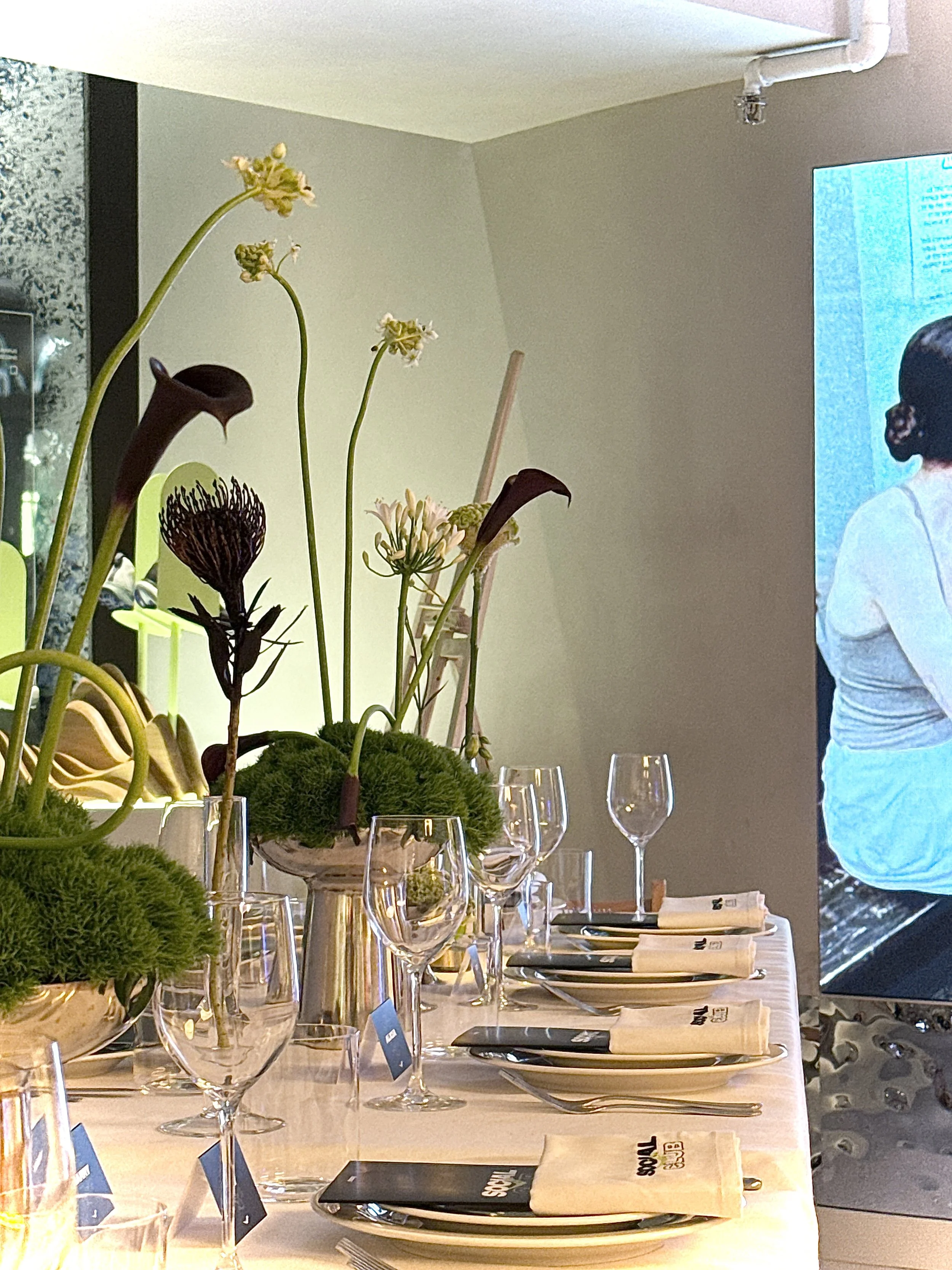 Elegant dining table setup with floral centerpieces, wine glasses, napkins, and cutlery, next to a large television screen, in a modern room.