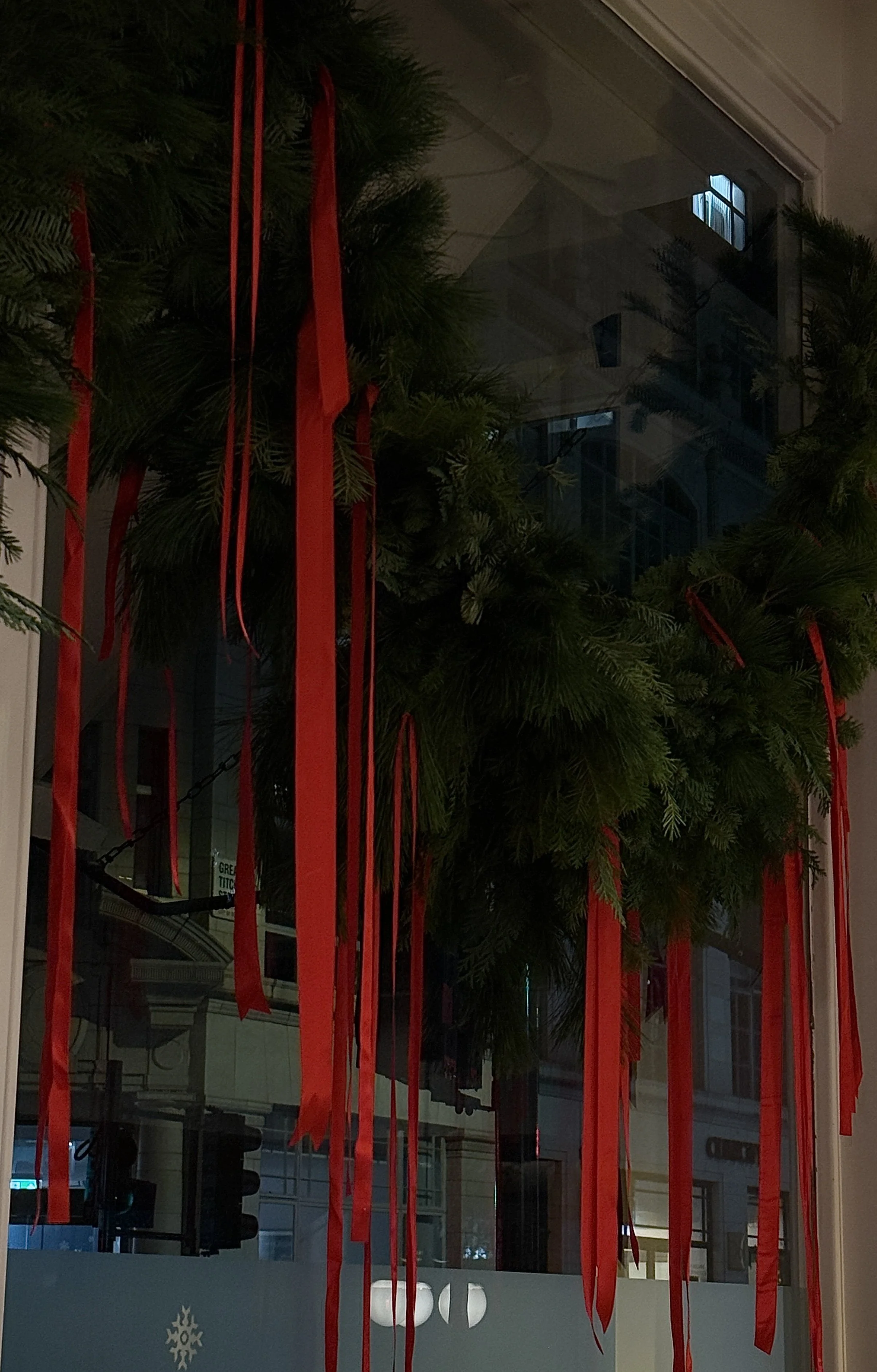 Christmas wreath with red ribbons hanging on a window, reflecting buildings and street lights outside.