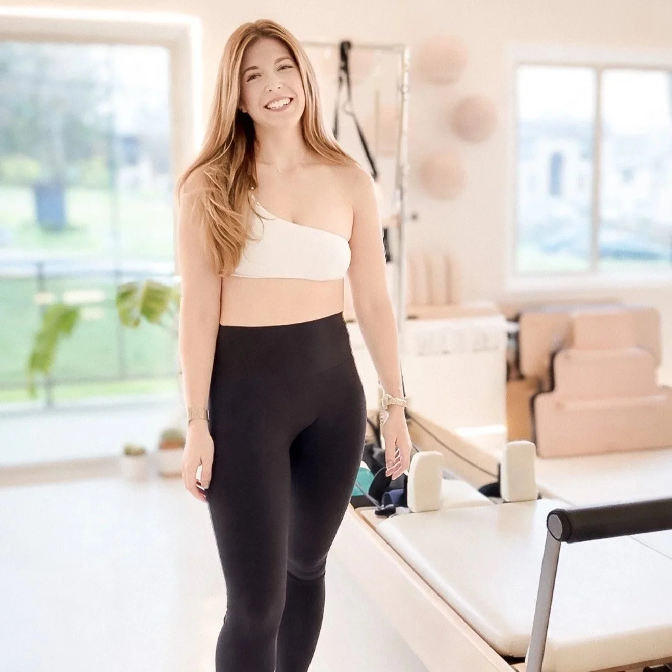 A woman with long red hair and a white one-shoulder top smiling in a bright, modern Pilates studio with natural light.