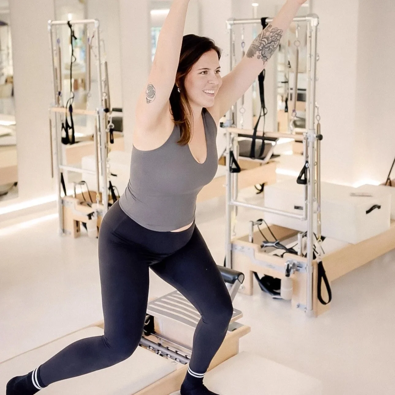 Woman in workout clothes exercising with arms raised in a Pilates studio with reformer equipment.