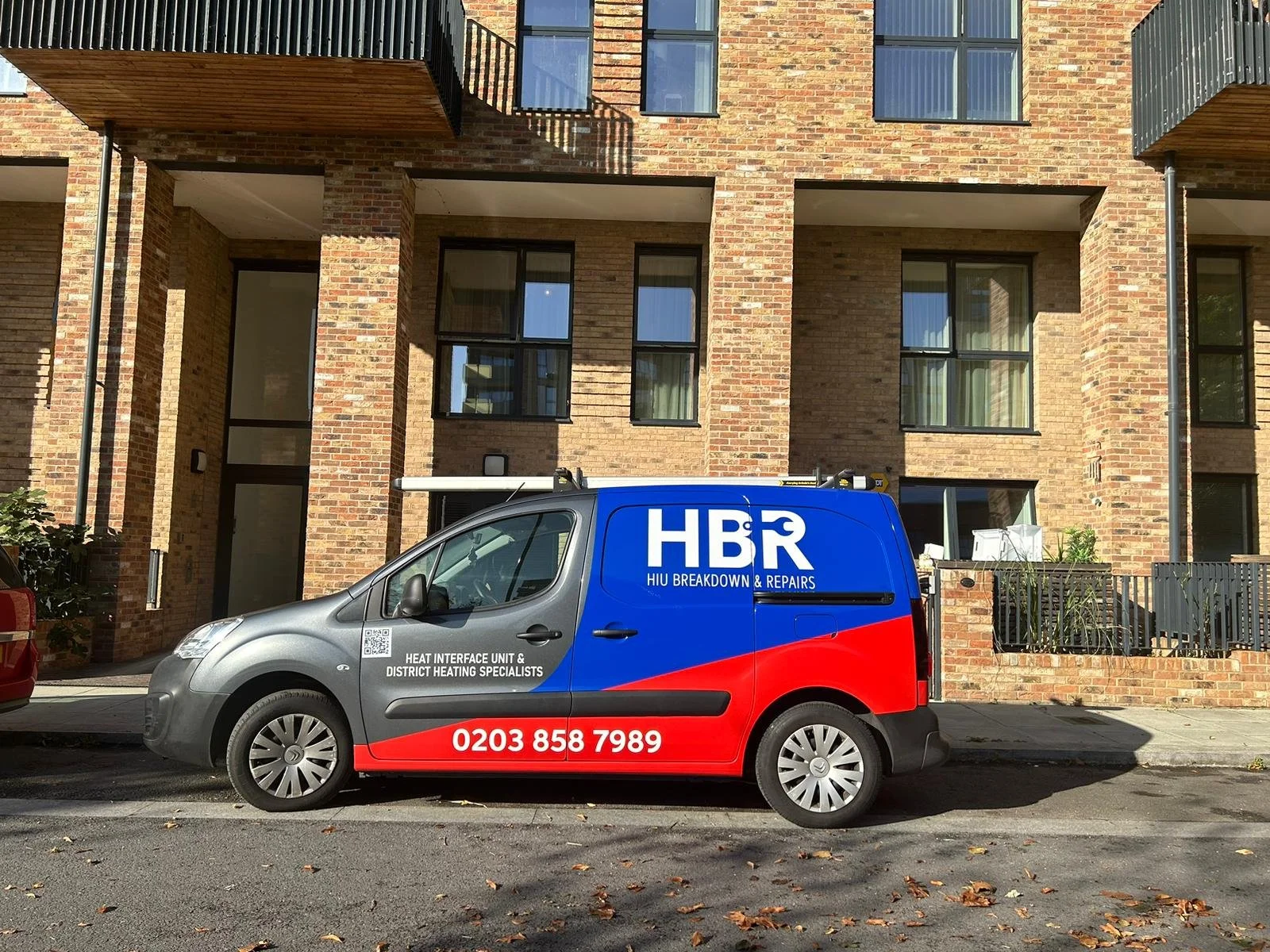 A repair van parked on a street in front of a modern brick apartment building. The van has the company name 'HBR' and contact information on its side.
