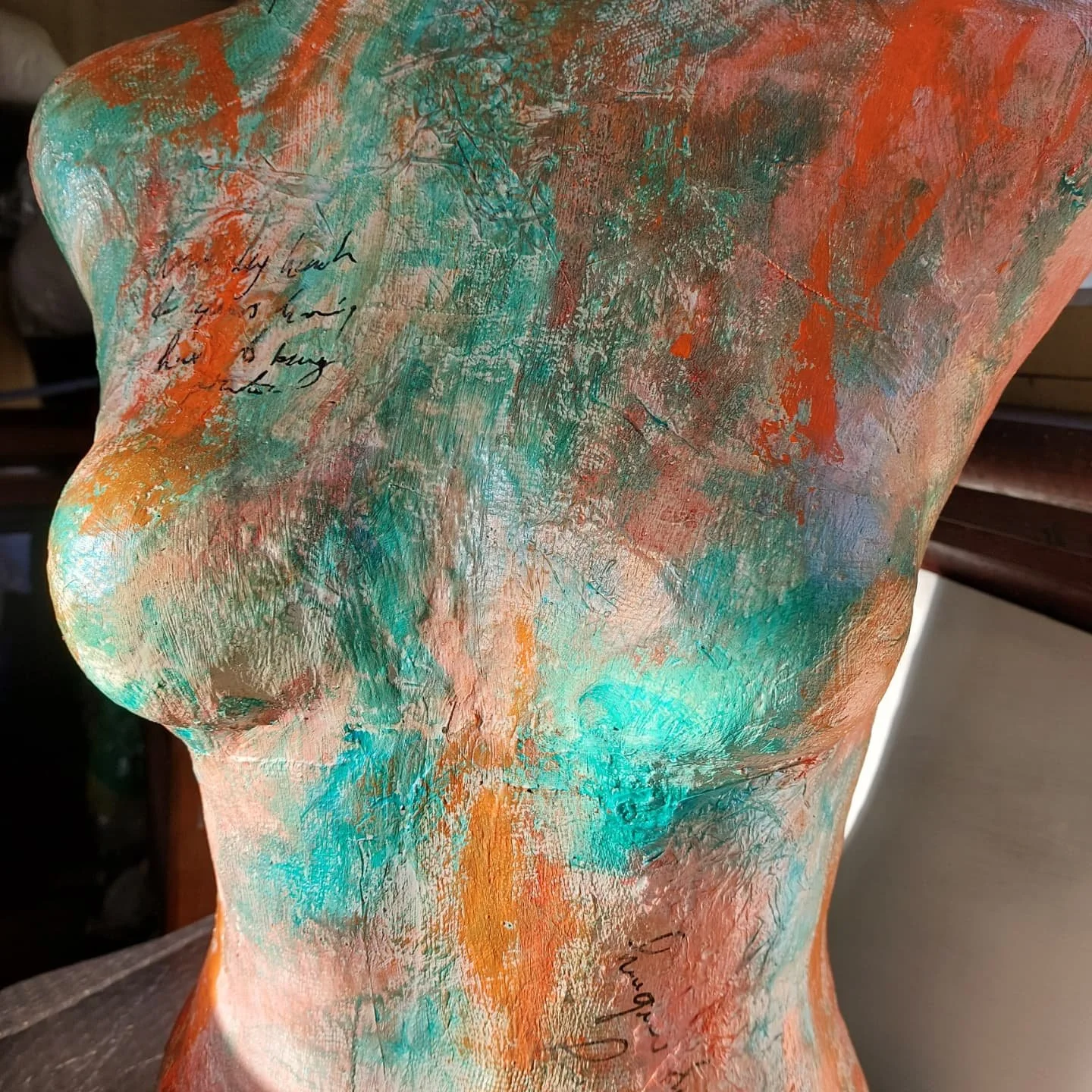 close-up of the torso after painting. The surface is now covered in energetic strokes of colour - vibrant coral, orange and teal green with metallic gold sheen catching the light. The texture is heavily layered with the earlier scribbled text beneath