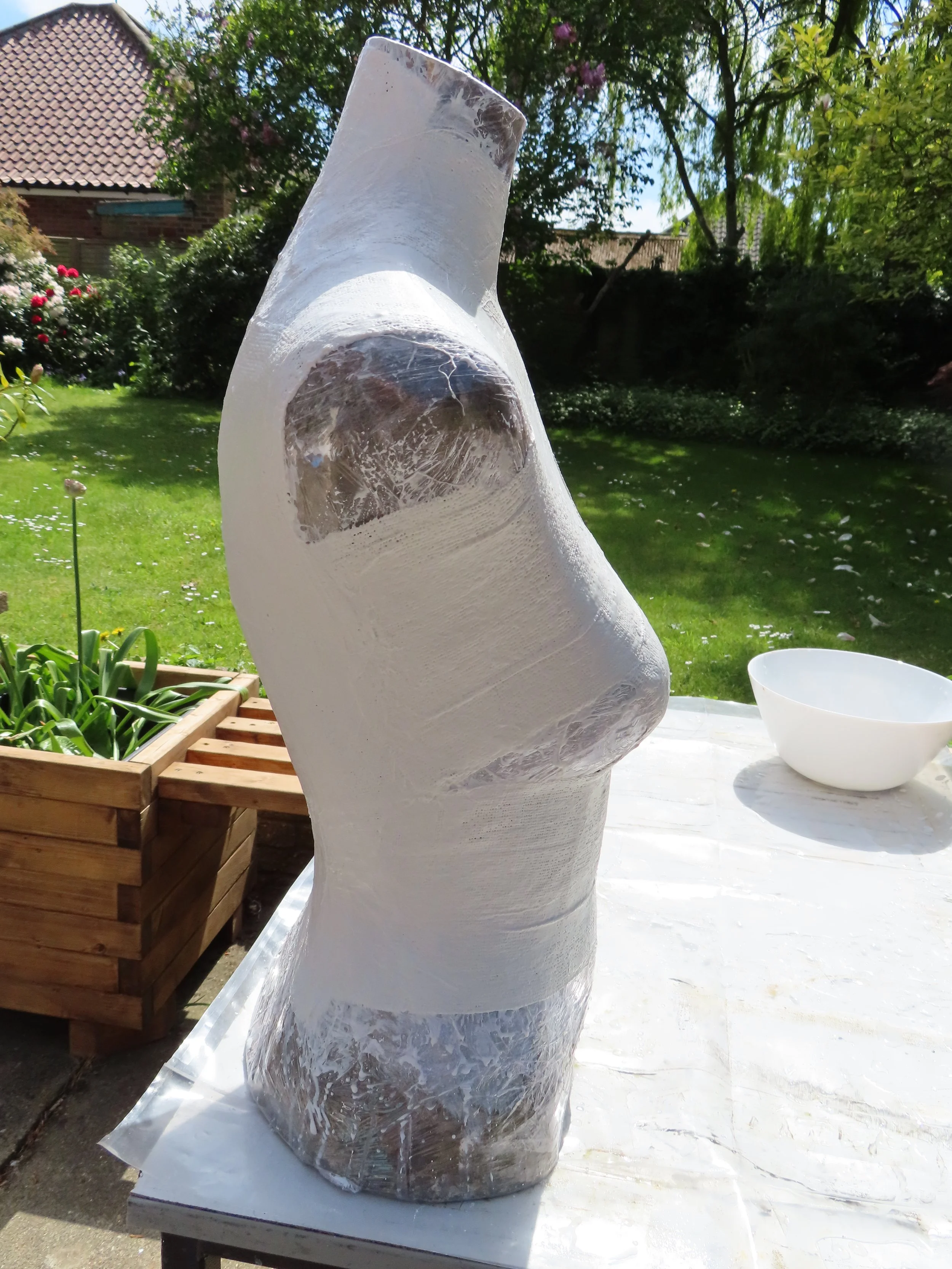 Side view of the sculpture’s early form: a white plaster torso cast made from modroc bandages, with cling film wrapped around the lower half. The unfinished torso stands on a table in a garden with grass and trees behind, drying in the sun.