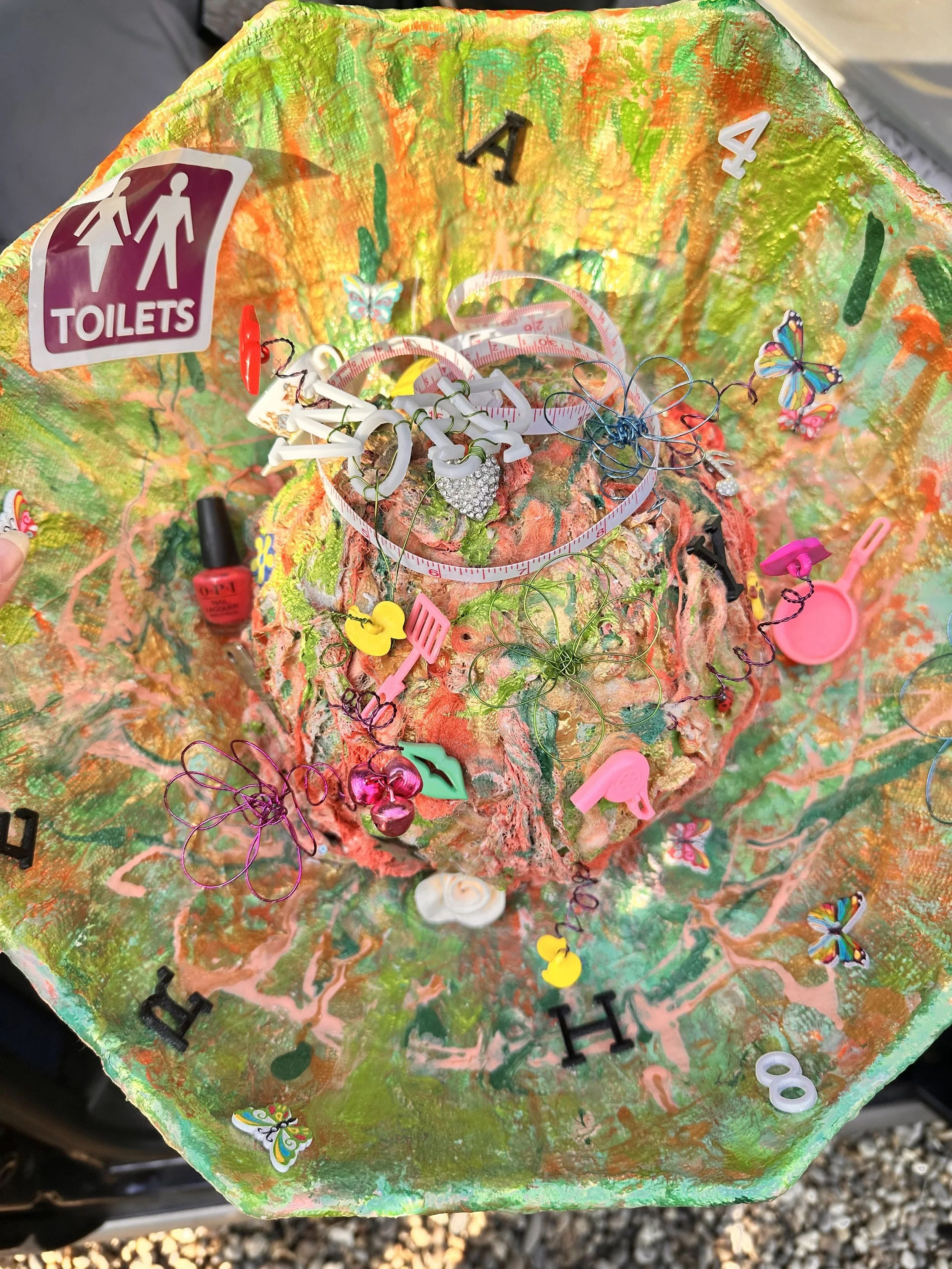 close-up into the sculpture’s brain bowl, showcasing a wild jumble of miniature objects embedded in colourful chaos. A coiled pink measuring tape, plastic letters and numbers, a pair of neon pink lips, a tiny yellow rubber duck.