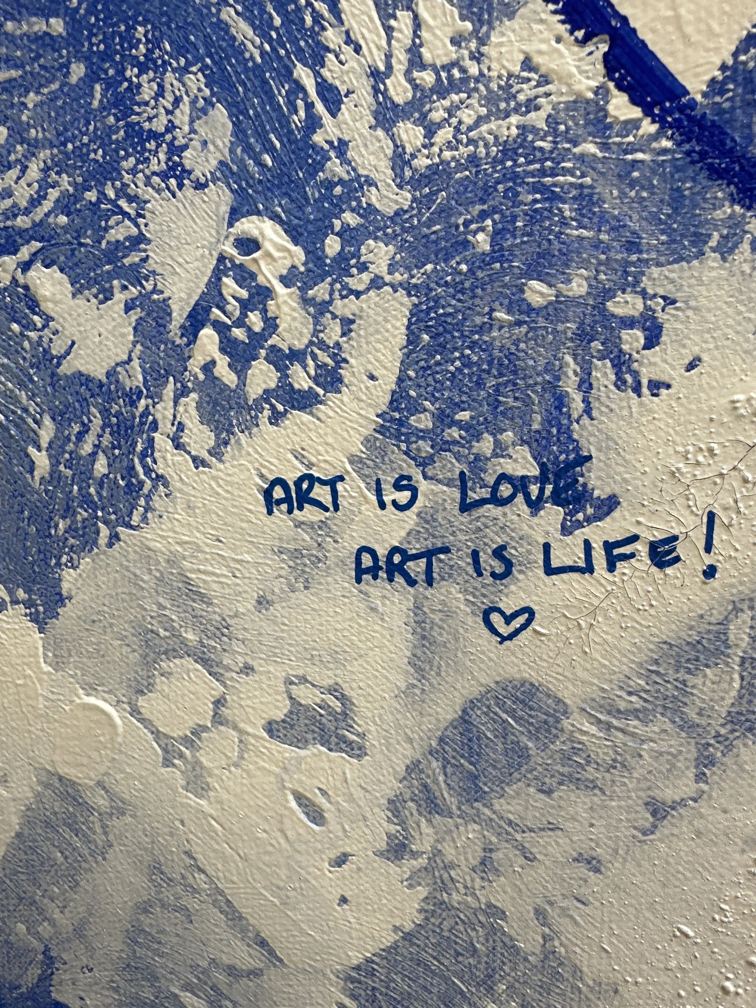 Close up of blue and white canvas painting, with textured paint and the phrase 'Art is Love, Art is Life!' written on the surface with a small heart drawn using a posca pen underneath the 'is'