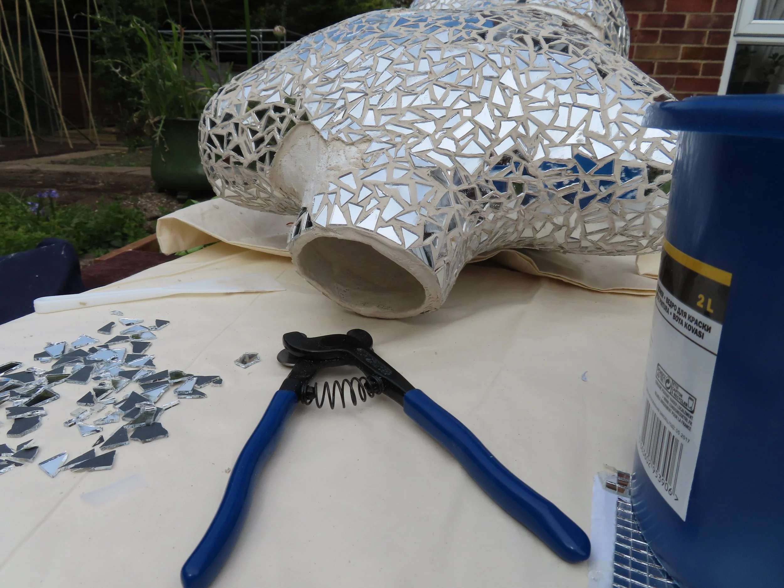 Close-up showing shards of mirror being glued to the bust sculpture of boudicca, with tools and broken glass nearby.