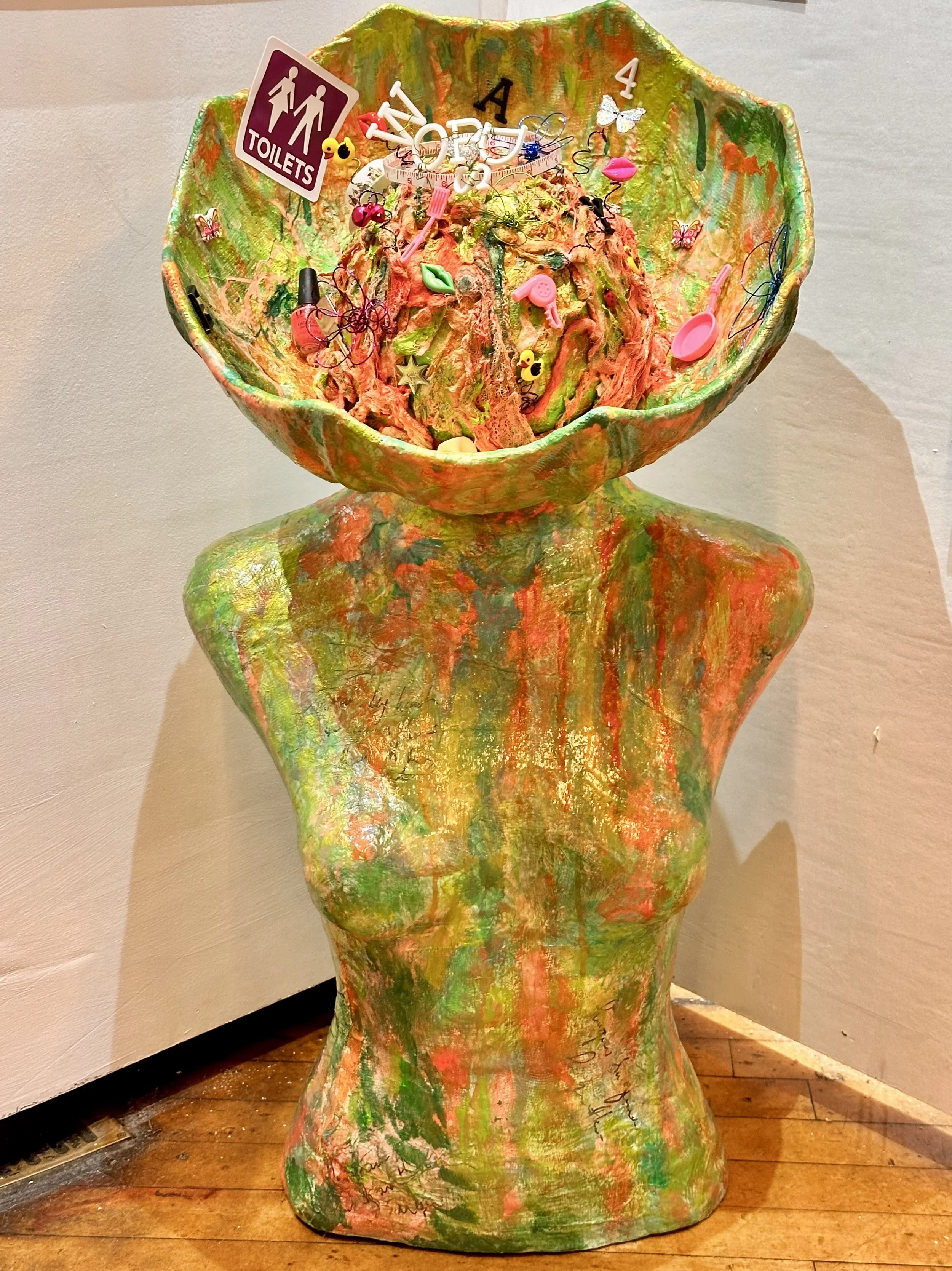 the large bowl-shaped brain opening, packed with its colourful assortment of tiny objects, and the textured torso gleams with green, orange and gold hues. The sculpture stands proudly upright on a white background