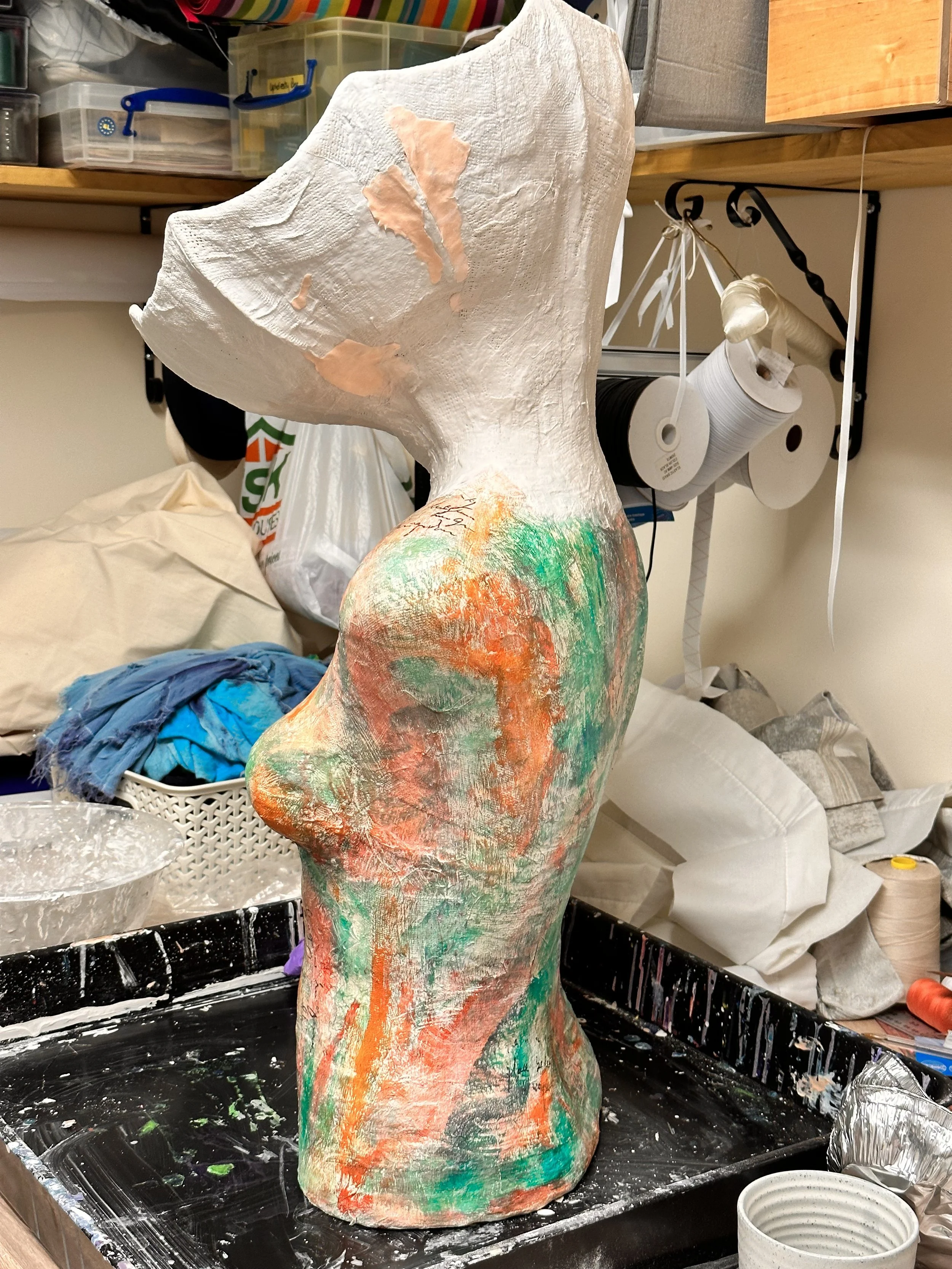 Profile view of the sculpture meno brin progress, taken in the studio. The torso is fully painted in mottled greens and oranges, but now an oversized bowl-like head has been added on top. The new “brain” section is still plain white and empty inside.