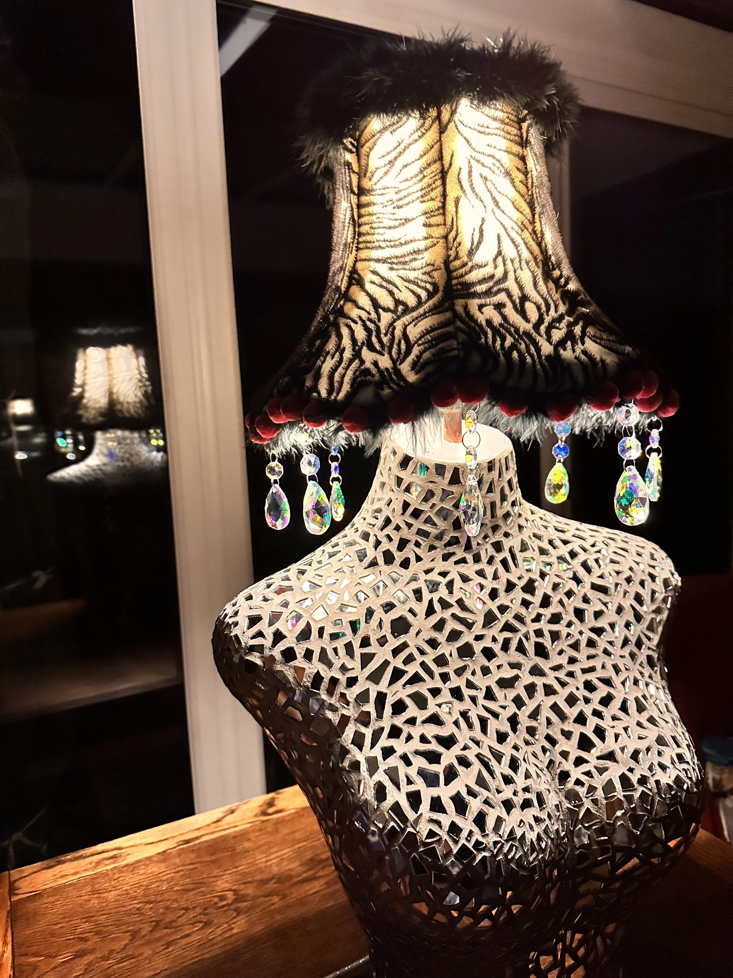 The sculpture of boudicca zebra lamp illuminated in a dark room, casting shimmering light through her mirrored torso and fabric shade.. rainbow aurora borealis crystals hang from the bottom of the shade which has black marabou feather pink pompoms