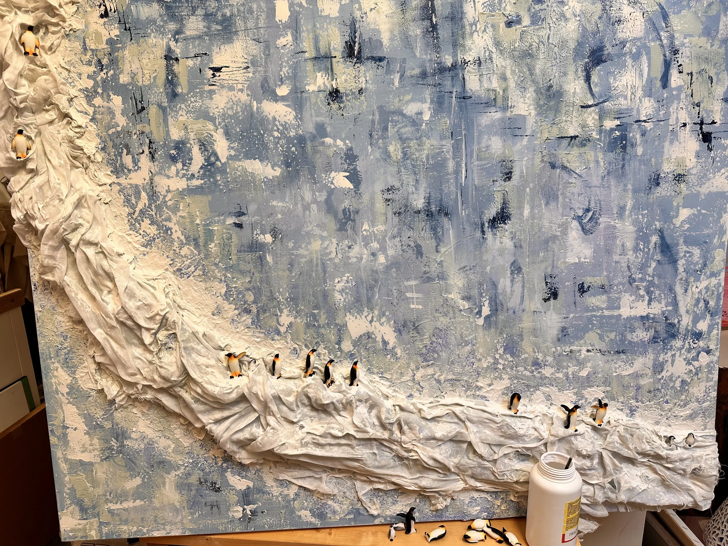Miniature penguins and chicks are being placed carefully along the muslin ledge, mid-composition. the blue white and green painted background is fully visible. the painting and some of the penguins sit on a wooden table