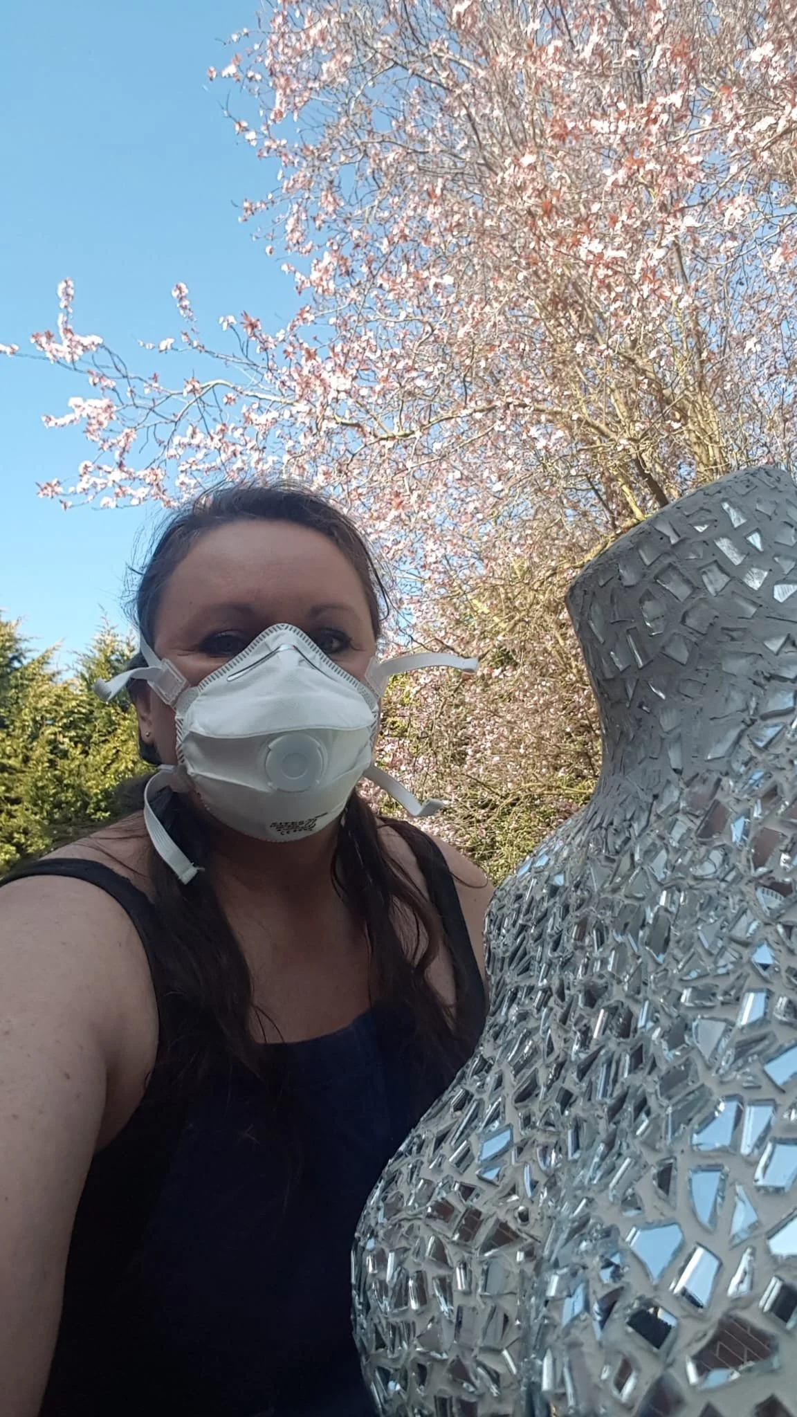 deborah phillips wearing a safety mask whilst grouting the sculpture torso of boudicca. close up of the mirror section of the chest and neck. magnolia tree in the background