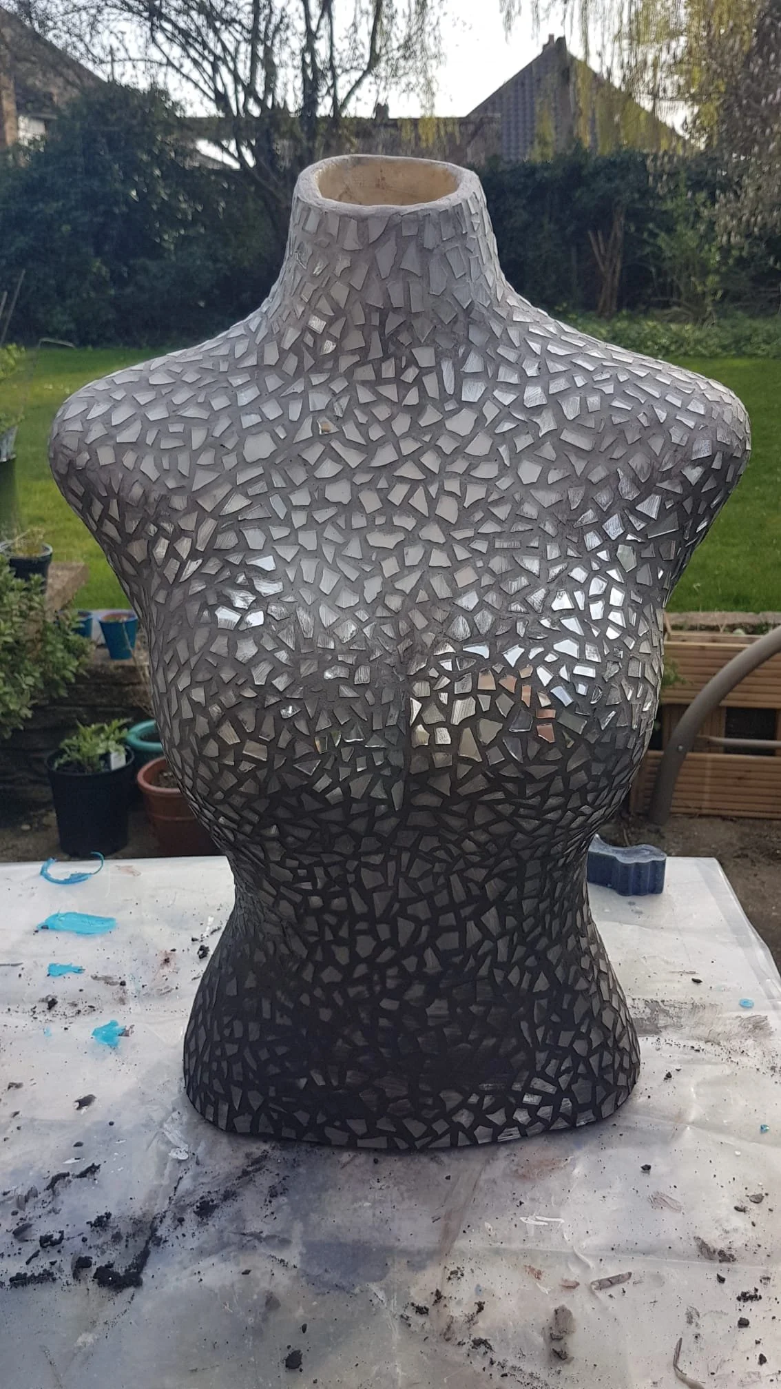 boudicca sculpture bust during construction having just been grouted with dark grey grout at the bottom of the torso graduating up to light grey around the neck. sitting on a table in a garden with bushes and plant pots in the background