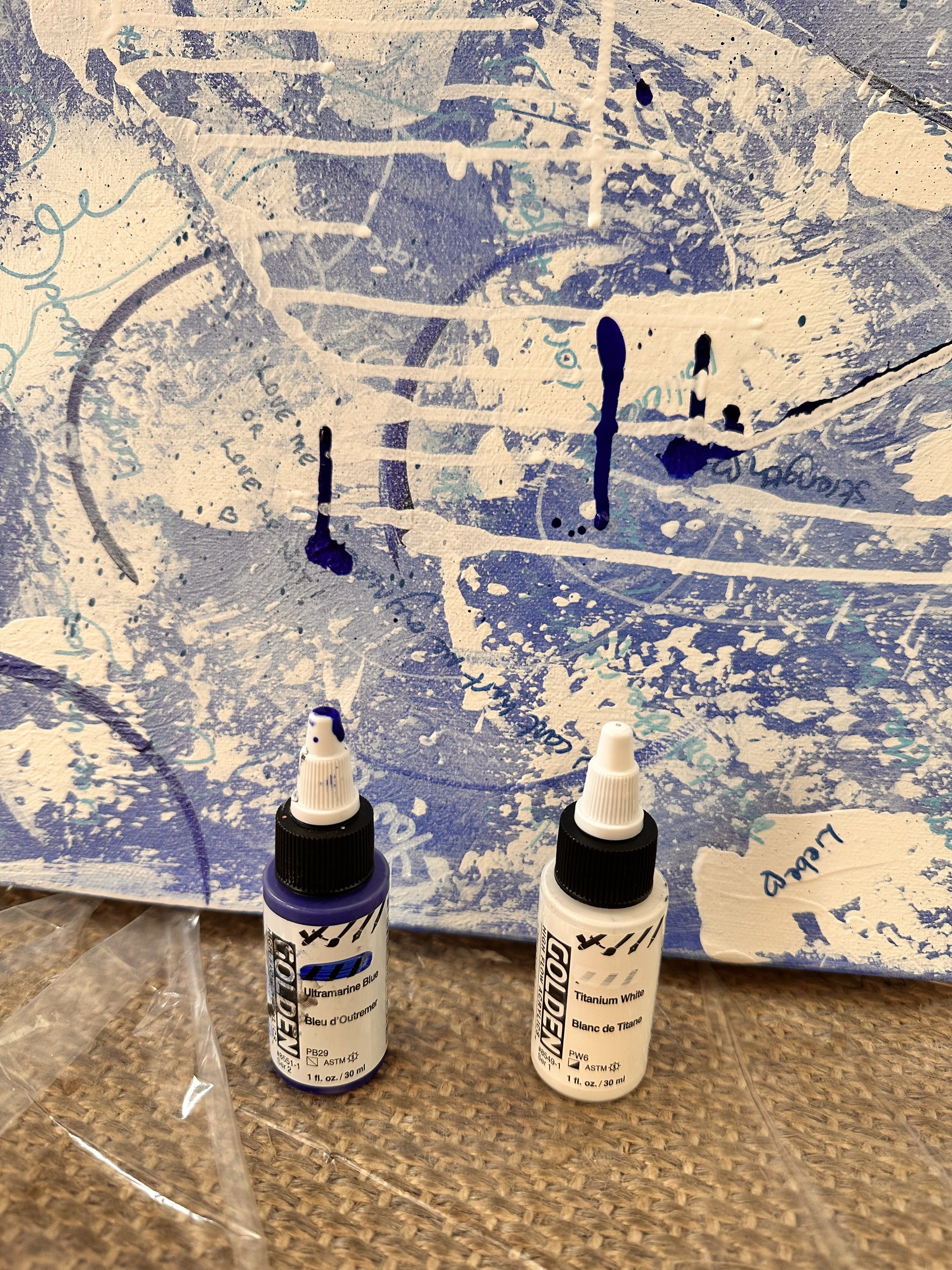 Section of a blue and white abstract painting with various 'healing' words and phrases written in blue and white ink, dribbles of ultramarine and titanium white 'golden' acrylic ink dripped over canvas, the bottles stand in front of the canvas