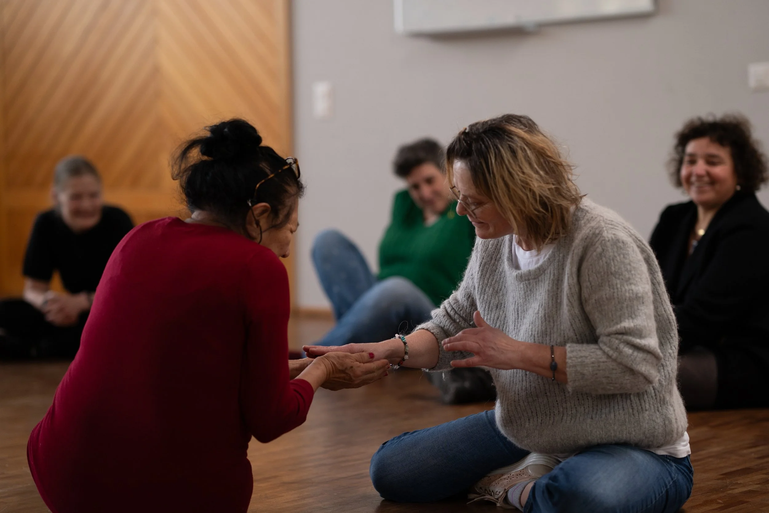 Deux femmes échangent un geste convivial, assises sur le sol, dans une pièce de style intérieur, probablement lors d'une réunion ou d'une activité de groupe, avec d'autres personnes souriantes en arrière-plan.