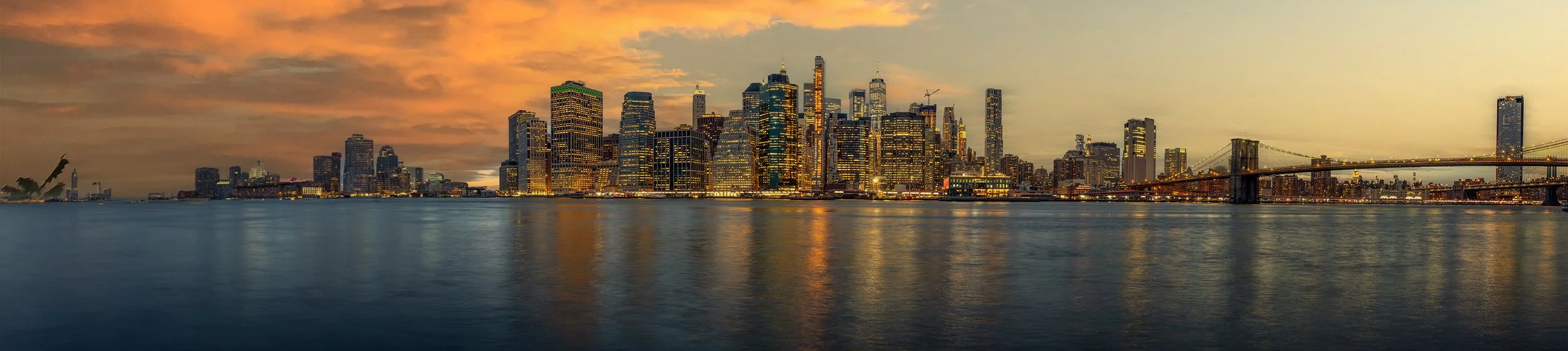 A panoramic view of a city skyline at sunset, with many illuminated skyscrapers reflecting on the water in the foreground and a bridge on the right side.