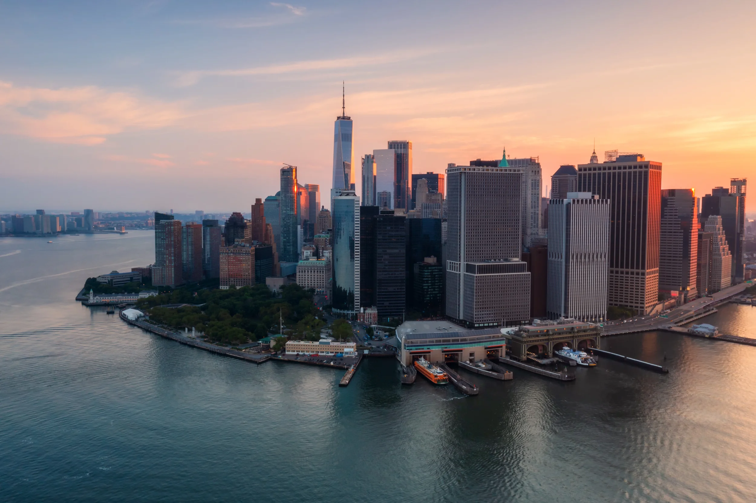 Aerial view of New York City skyline at sunset, featuring tall skyscrapers along the waterfront with calm water in the foreground.