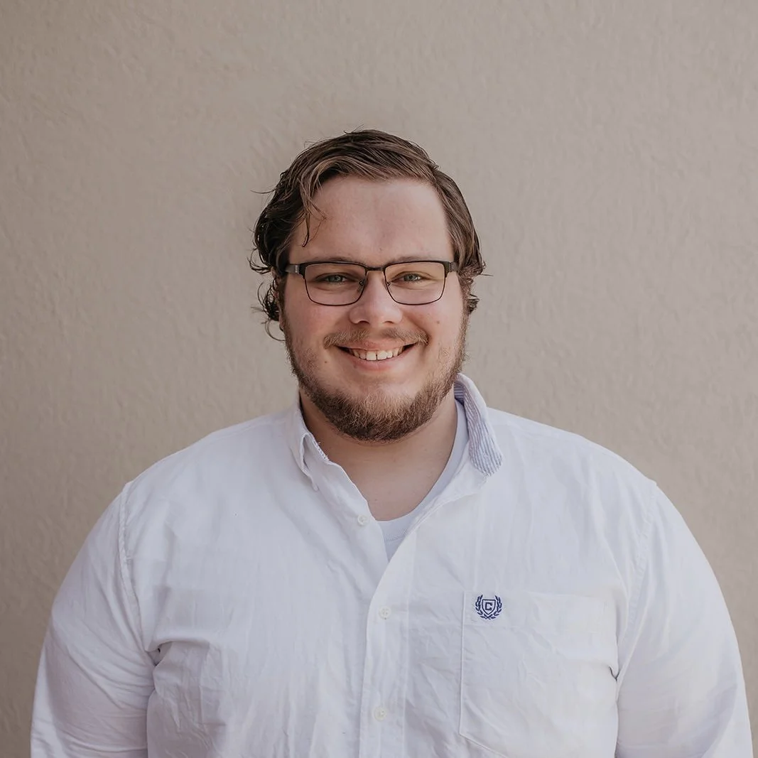 A smiling man with glasses and a beard wearing a white button-up shirt standing against a beige wall.