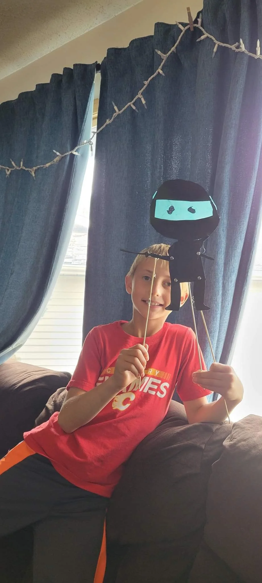 A boy holding a homemade ninja puppet, made from black and blue materials, in front of dark curtains with string lights above.